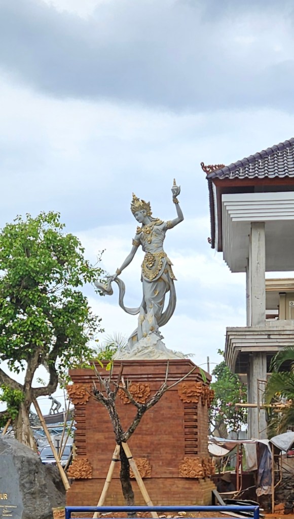 A detailed statue of a deity holding a flower, situated on a carved pedestal, with a backdrop of cloudy skies and nearby greenery.