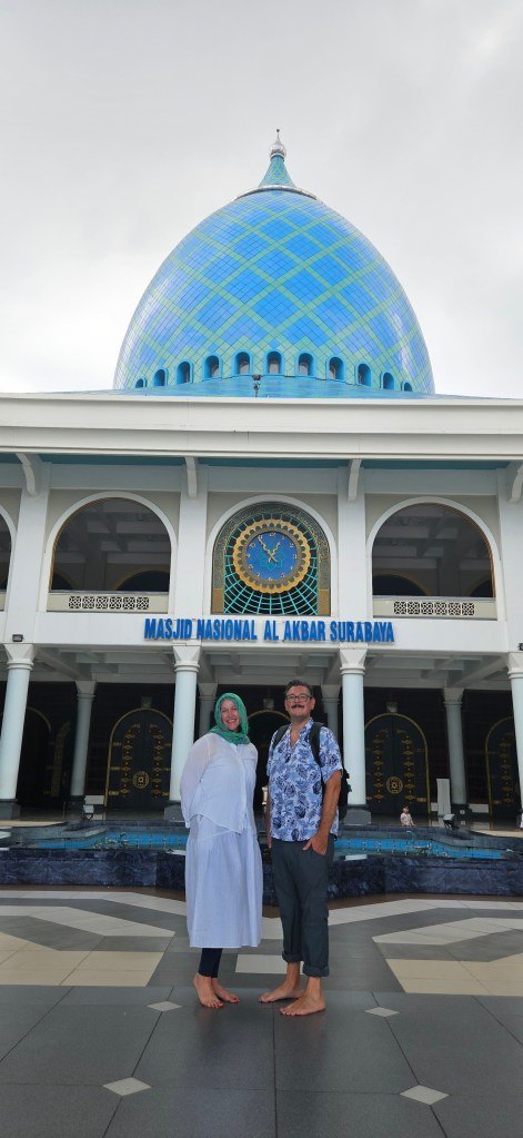 A woman in a white dress and green hijab and a man in a patterned shirt stand together in front of the Al Akbar National Mosque in Surabaya, Indonesia. The mosque features a large blue dome with diamond patterns and a clock above the entrance.