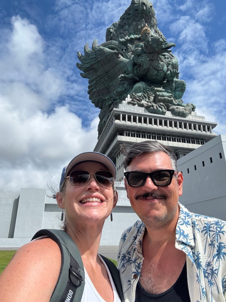 A smiling couple taking a selfie in front of a large statue, with cloudy skies above.