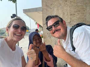 Three people taking a selfie outdoors, smiling and giving thumbs up. Two are wearing sunglasses, with a woman on the left and a man on the right. In the middle, a woman in a hijab also gives a thumbs up.