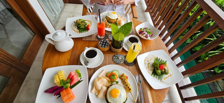 A beautifully arranged breakfast table featuring various dishes, including eggs, fruit, and salad, with drinks like coffee and juice, set on a wooden table near a lush garden.