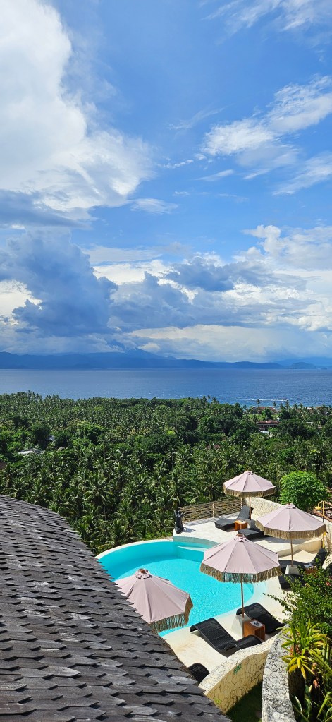 A scenic view of a luxury resort featuring a swimming pool with umbrellas, surrounded by lush palm trees and overlooking the ocean under a partly cloudy sky.