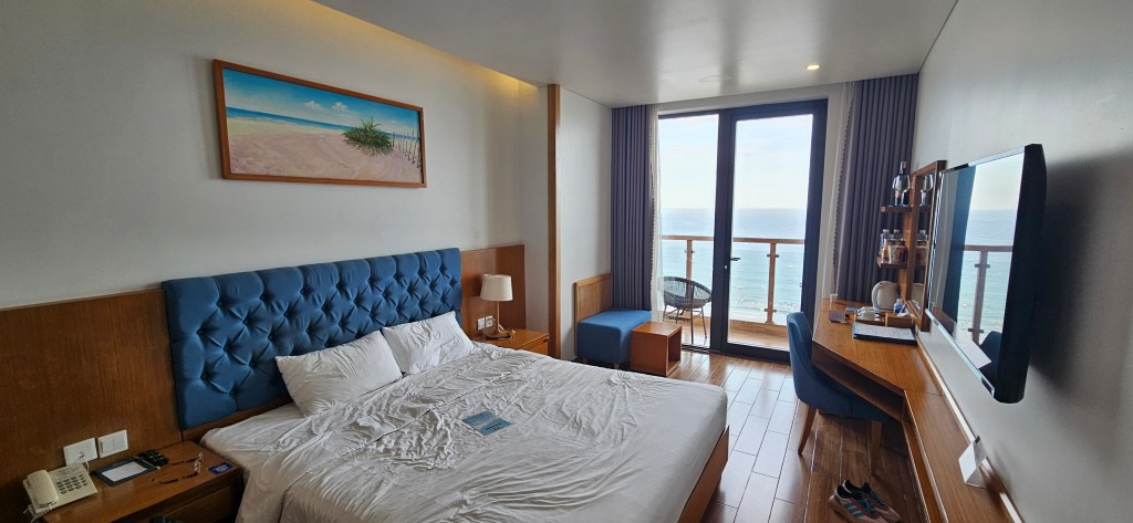 Interior of a hotel room featuring a king-sized bed with a tufted headboard, a wooden desk with a chair, and a balcony with ocean views.