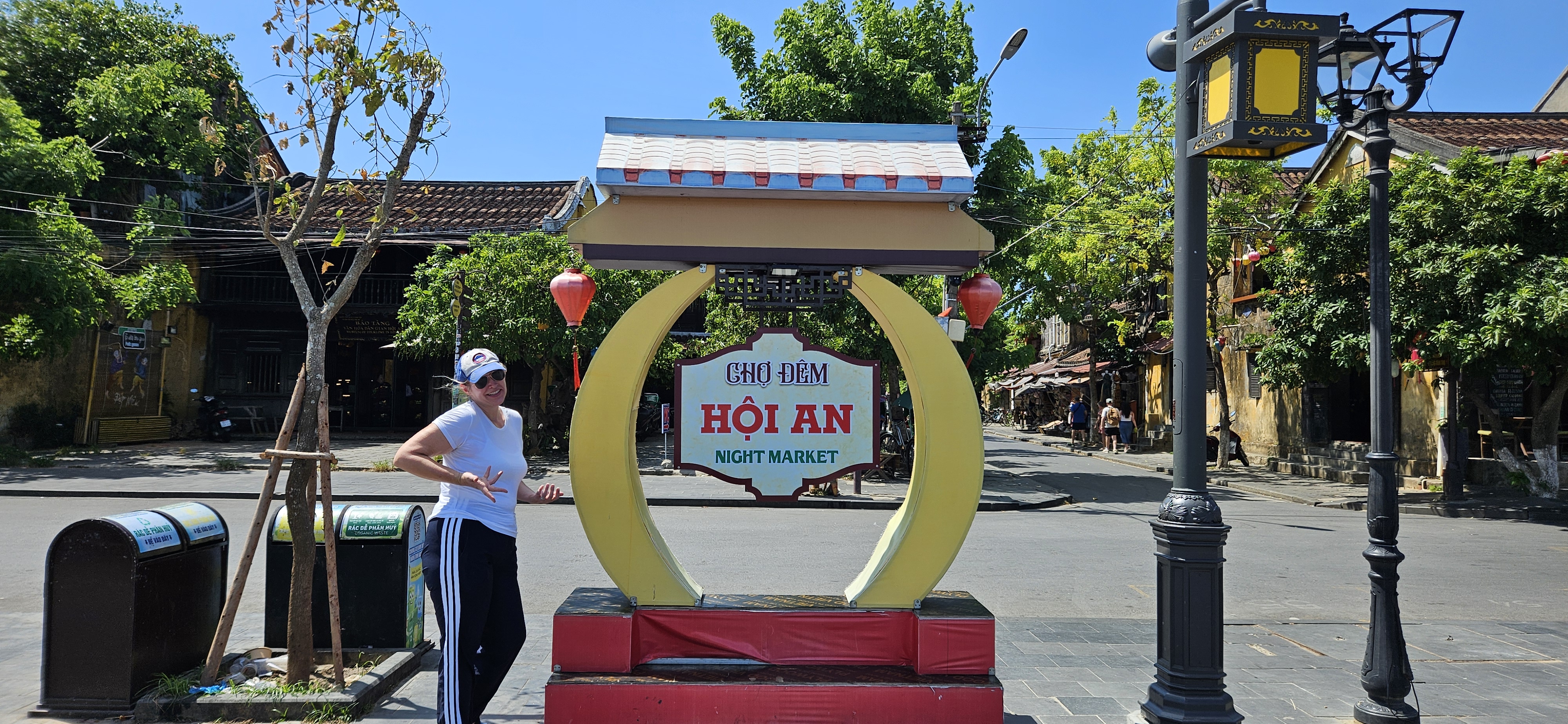 A person standing next to a colorful sign for the Hoi An Night Market, with shops and trees in the background under a clear blue sky.