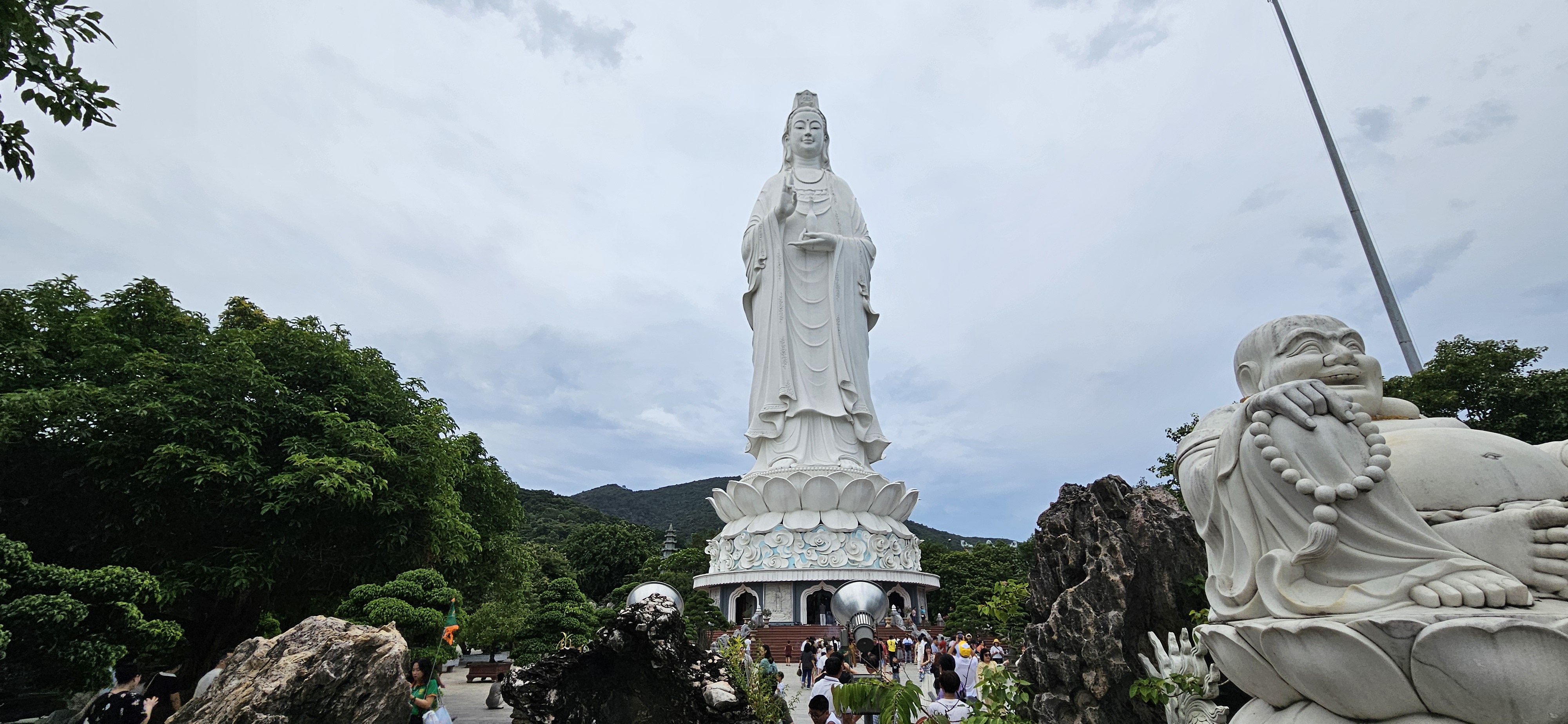 A tall white statue of a goddess standing on a lotus pedestal, surrounded by greenery and visitors, with a smaller statue of a smiling figure nearby against a cloudy sky.