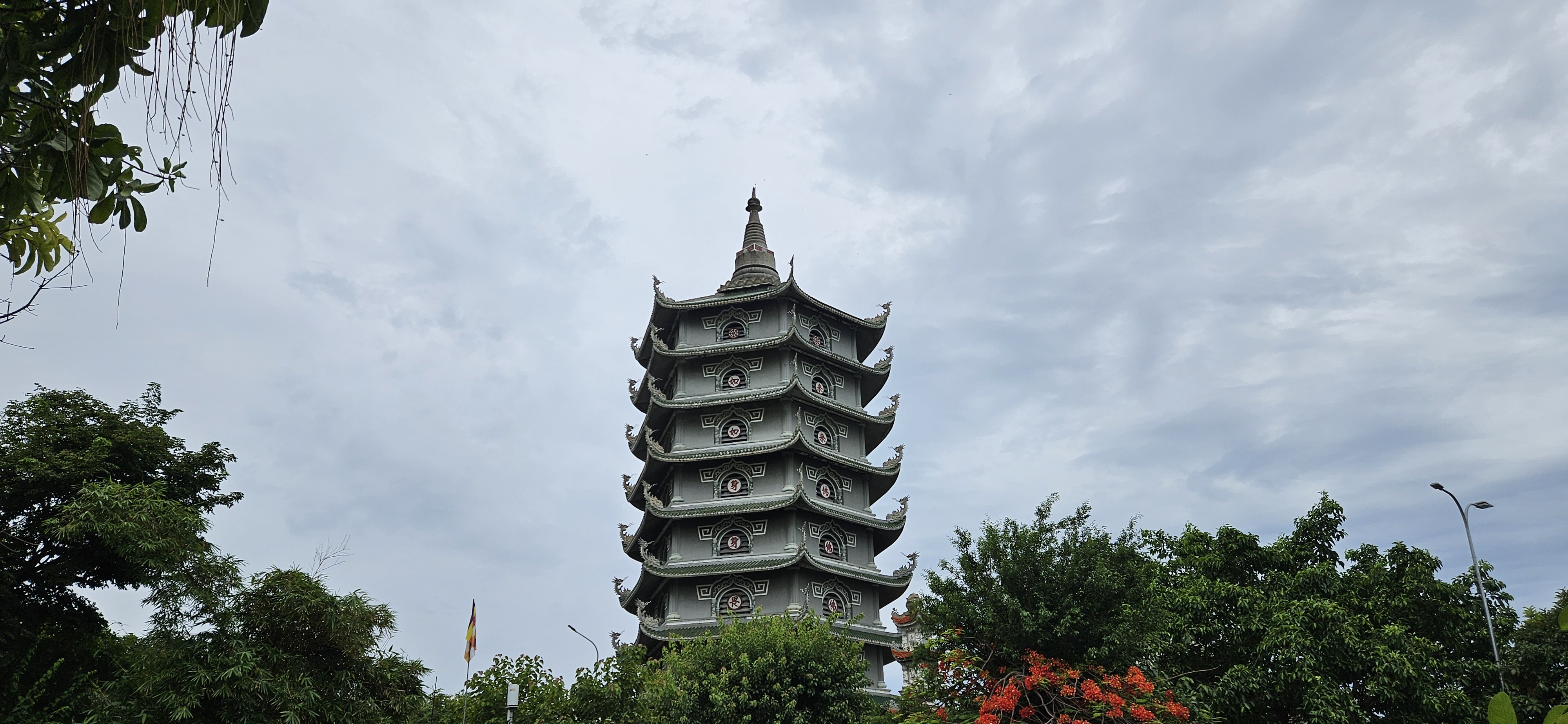 A tall, ornate pagoda surrounded by trees and colorful flowers under a cloudy sky.