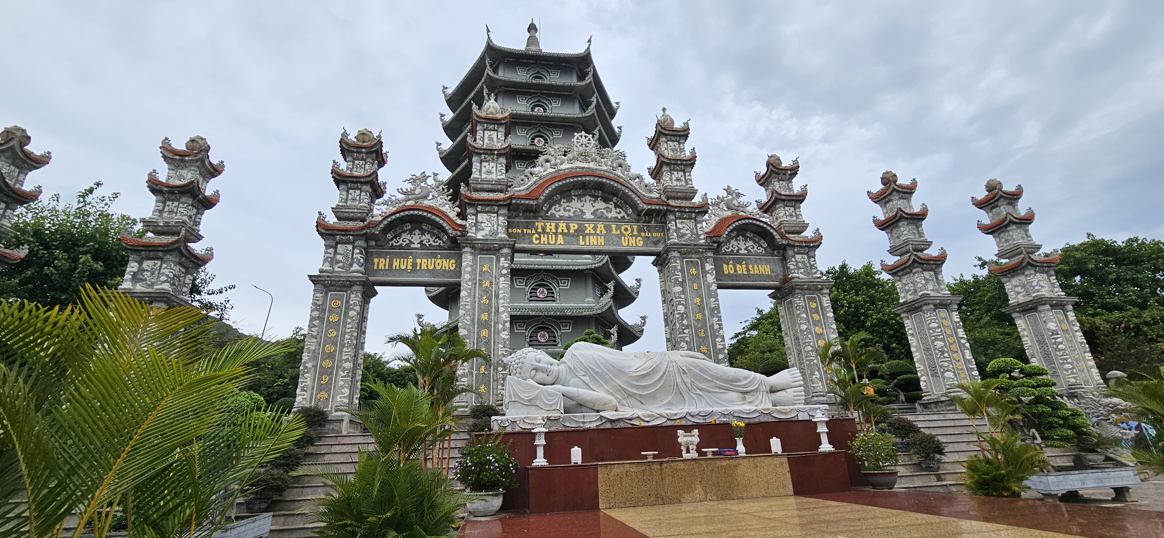 A pagoda-style structure with intricate architecture and decorative pillars, featuring a large reclining Buddha statue in a landscaped garden.