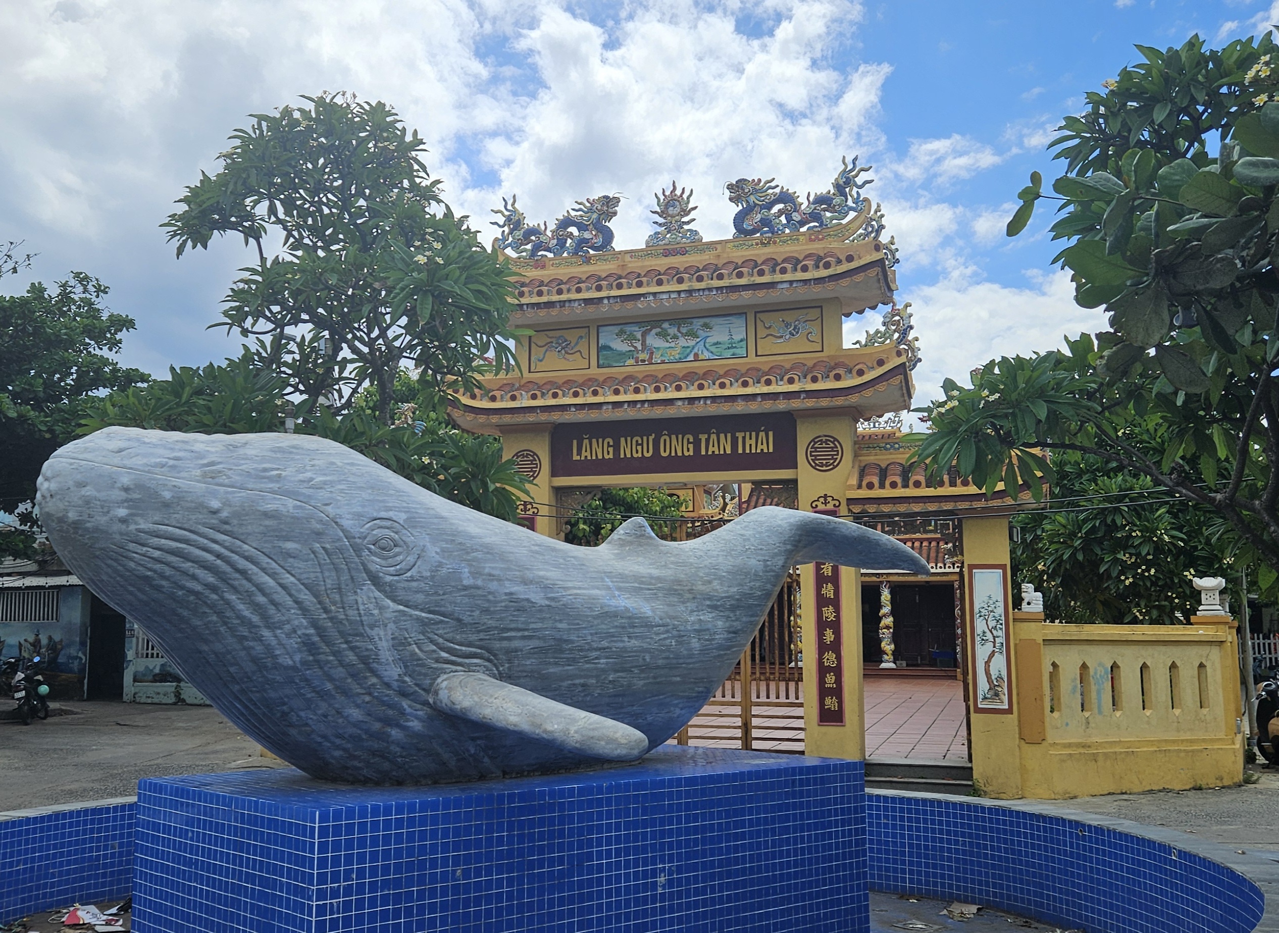 A large stone sculpture of a whale in front of a traditional ornate entrance gate, surrounded by trees and blue tiles.
