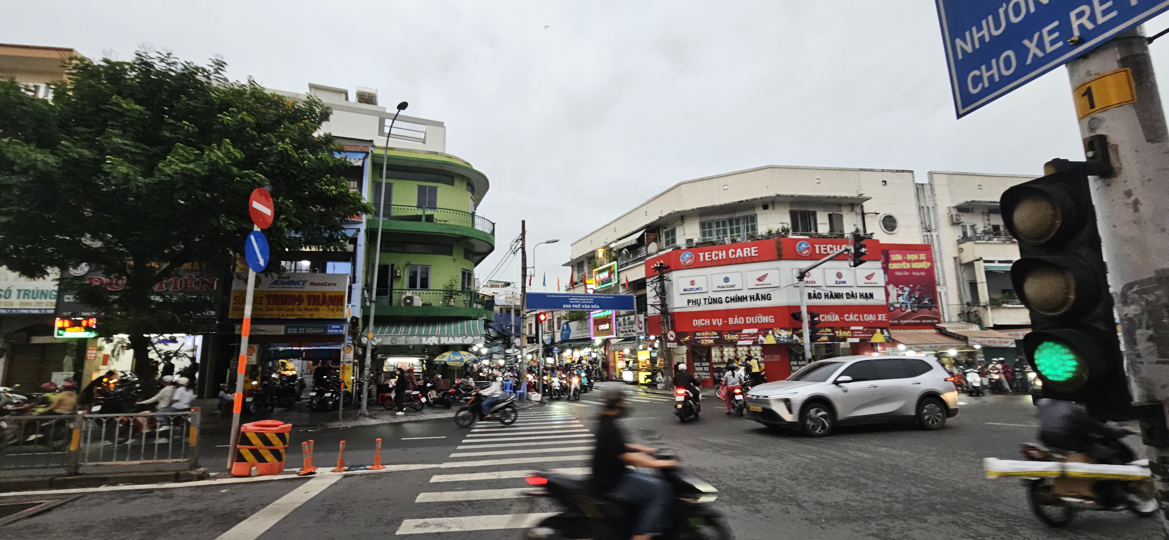 A busy urban intersection with traffic lights and multiple vehicles, including motorbikes and cars, surrounded by shops and buildings. The scene is set in the evening with cloudy skies.