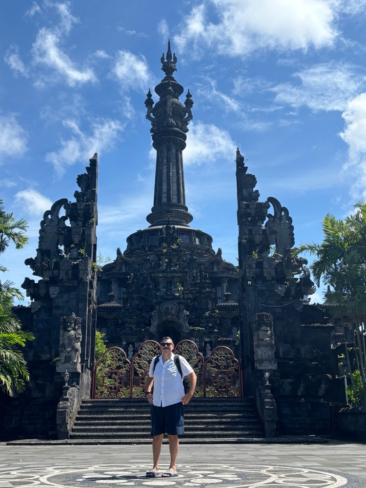A person standing in front of a large stone structure with intricate carvings and a tall spire, surrounded by greenery and under a clear blue sky.