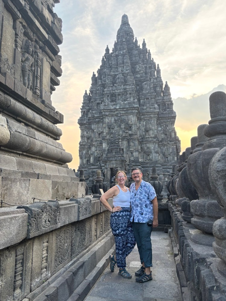 A couple posing for a photo in front of a large, intricate stone temple structure during sunset. The temple features detailed carvings and is surrounded by ancient stone features.