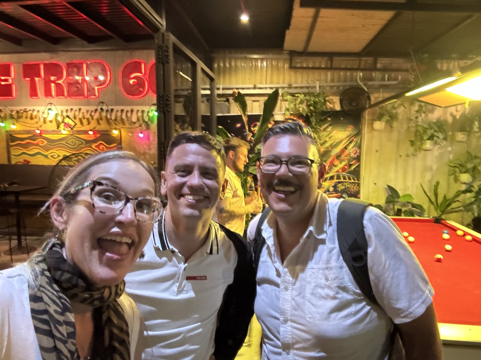 Three friends posing for a selfie in a lively bar with colorful lights and a pool table in the background.