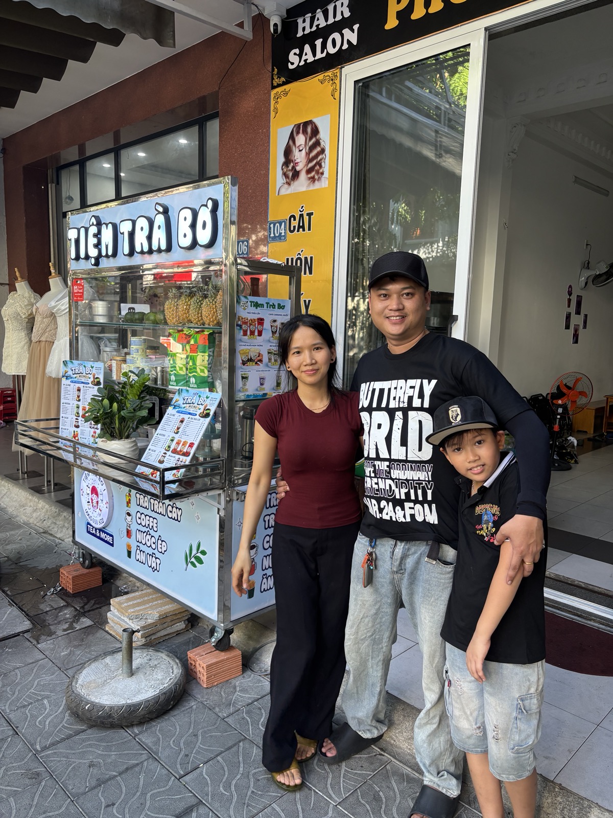 A family poses outside a tea shop with a colorful sign that reads 'Tiệm Trà Bồ'. They are standing on a sidewalk, with various items displayed in the shop's window and a hair salon sign visible above.