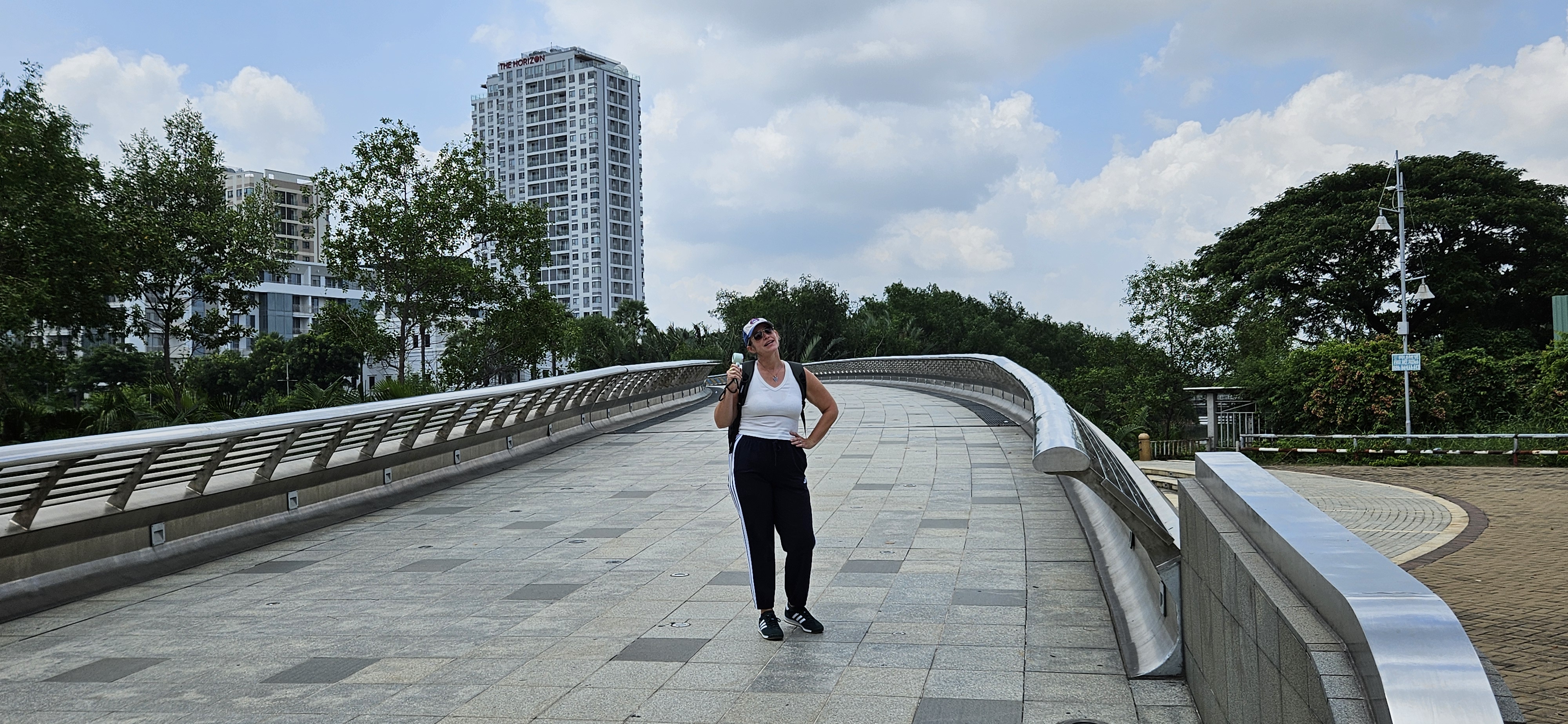 A woman stands on a modern bridge with a smile, wearing a white tank top and black pants, with buildings and greenery in the background.