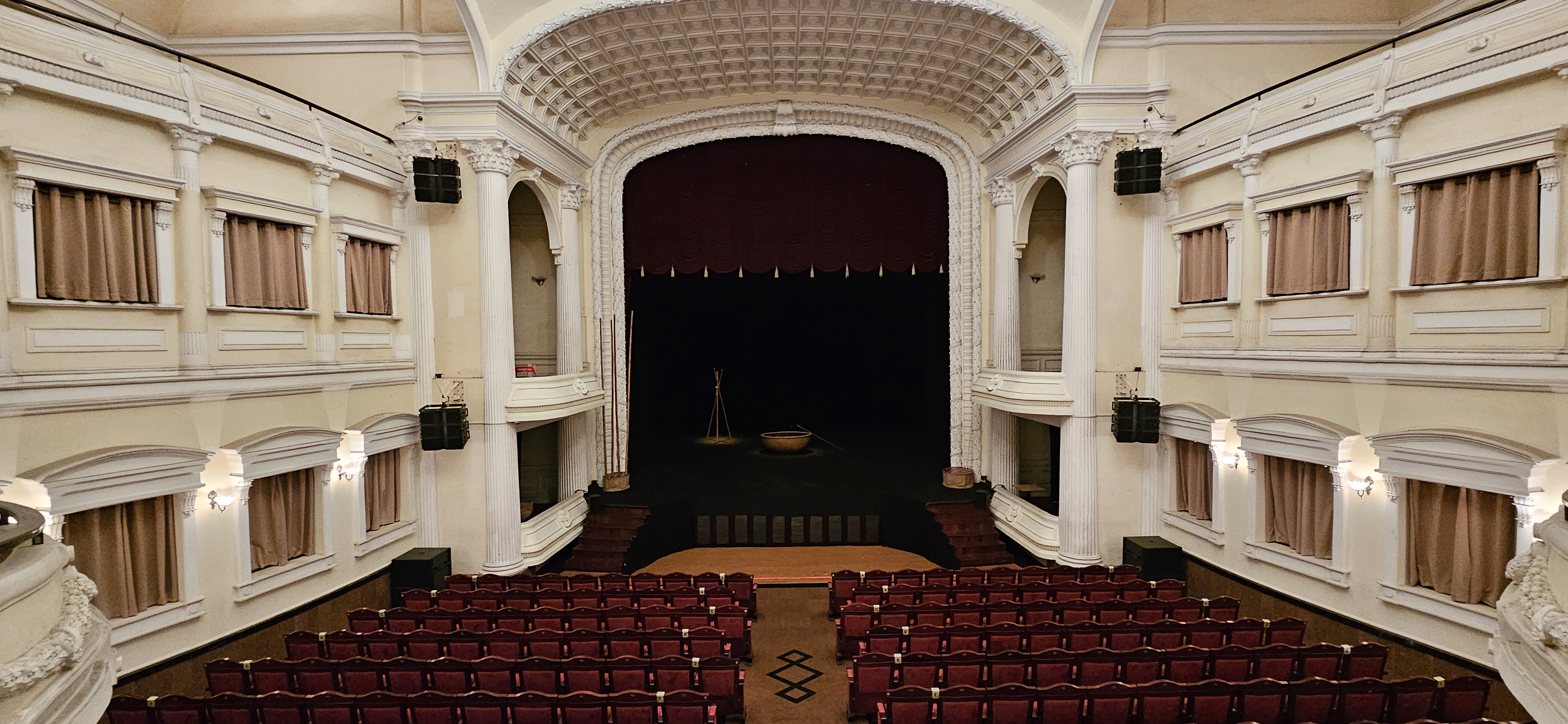 Interior view of a theater with red seats, a stage with dark curtains, and decorative architectural details.