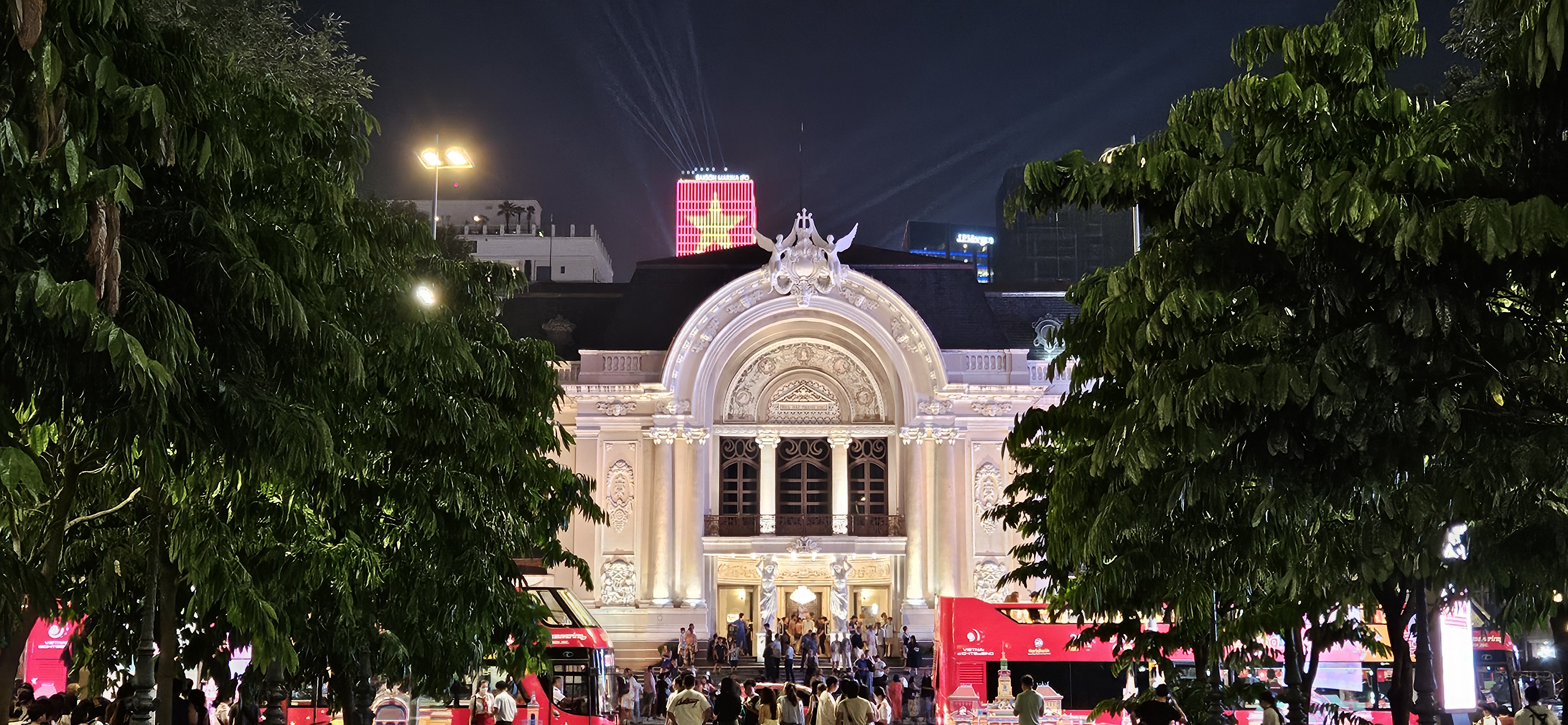 Night view of a historic building illuminated with lights, surrounded by green trees and bustling crowds, with a tall building featuring a star motif in the background.