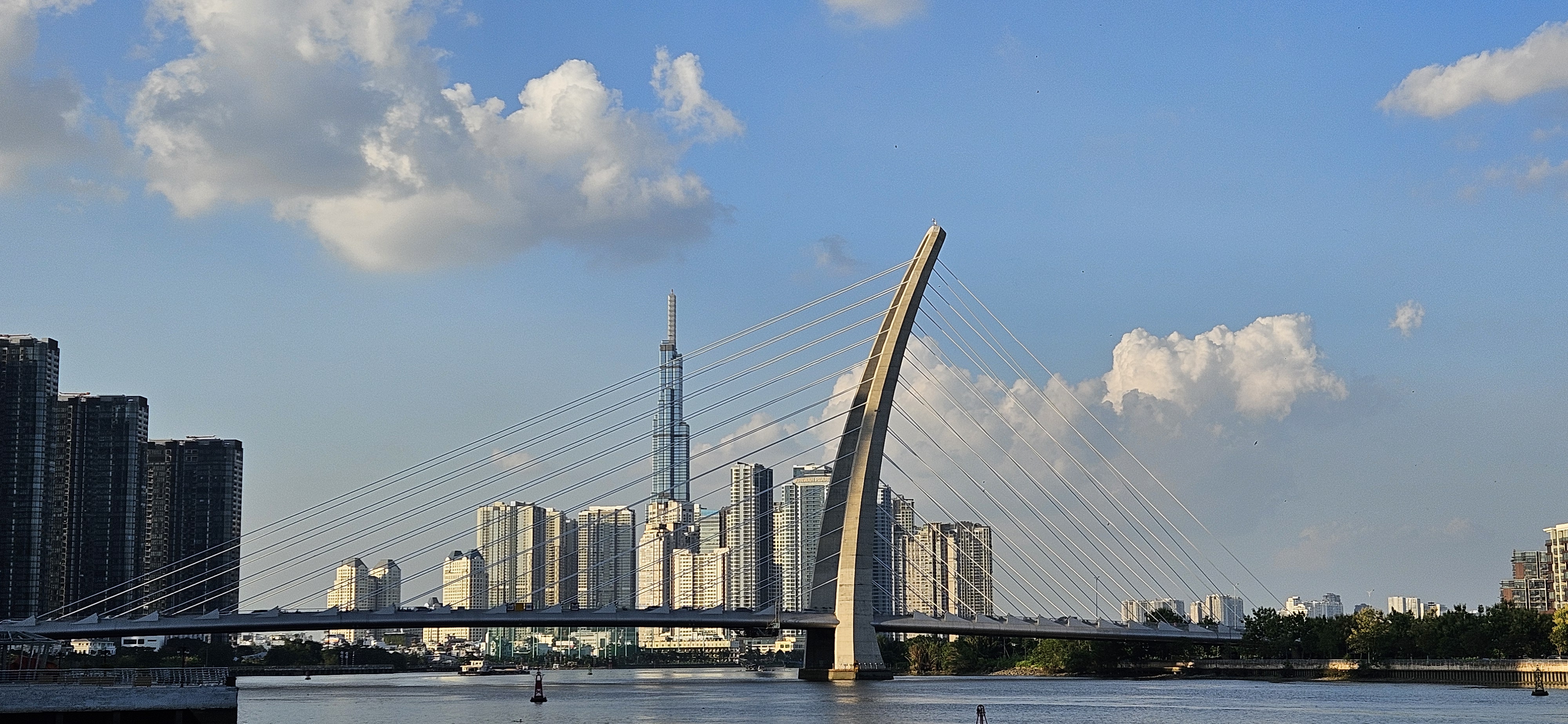 A modern cable-stayed bridge spans a river with a city skyline in the background, featuring tall buildings and partly cloudy skies.