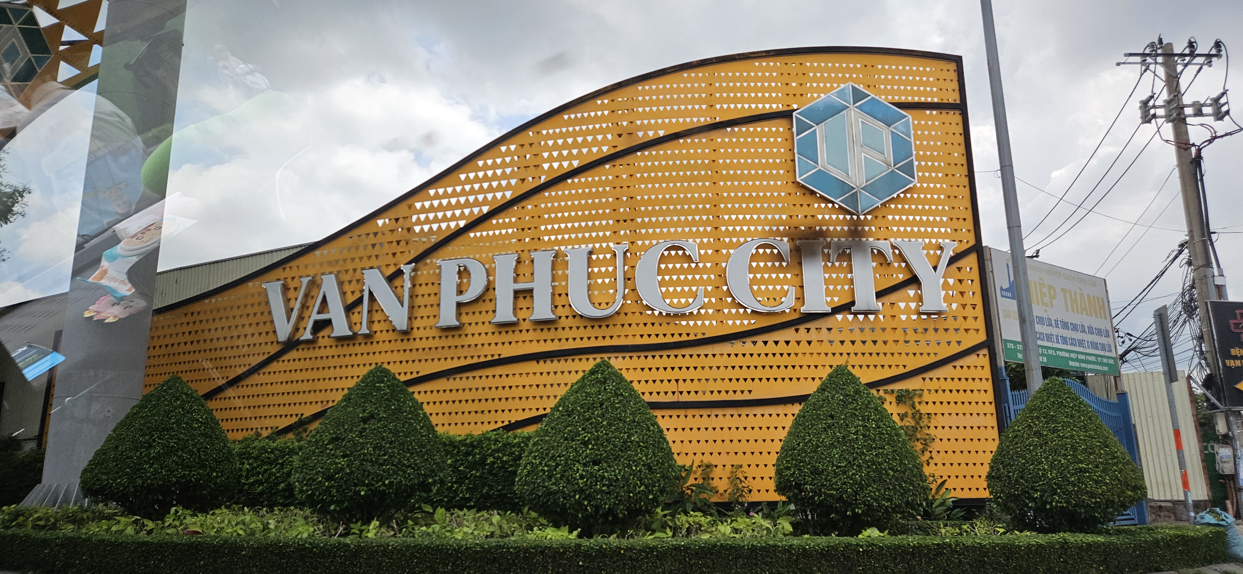 Signage for Van Phuc City, featuring a modern design and orange background, surrounded by manicured hedges and outlined against a cloudy sky.