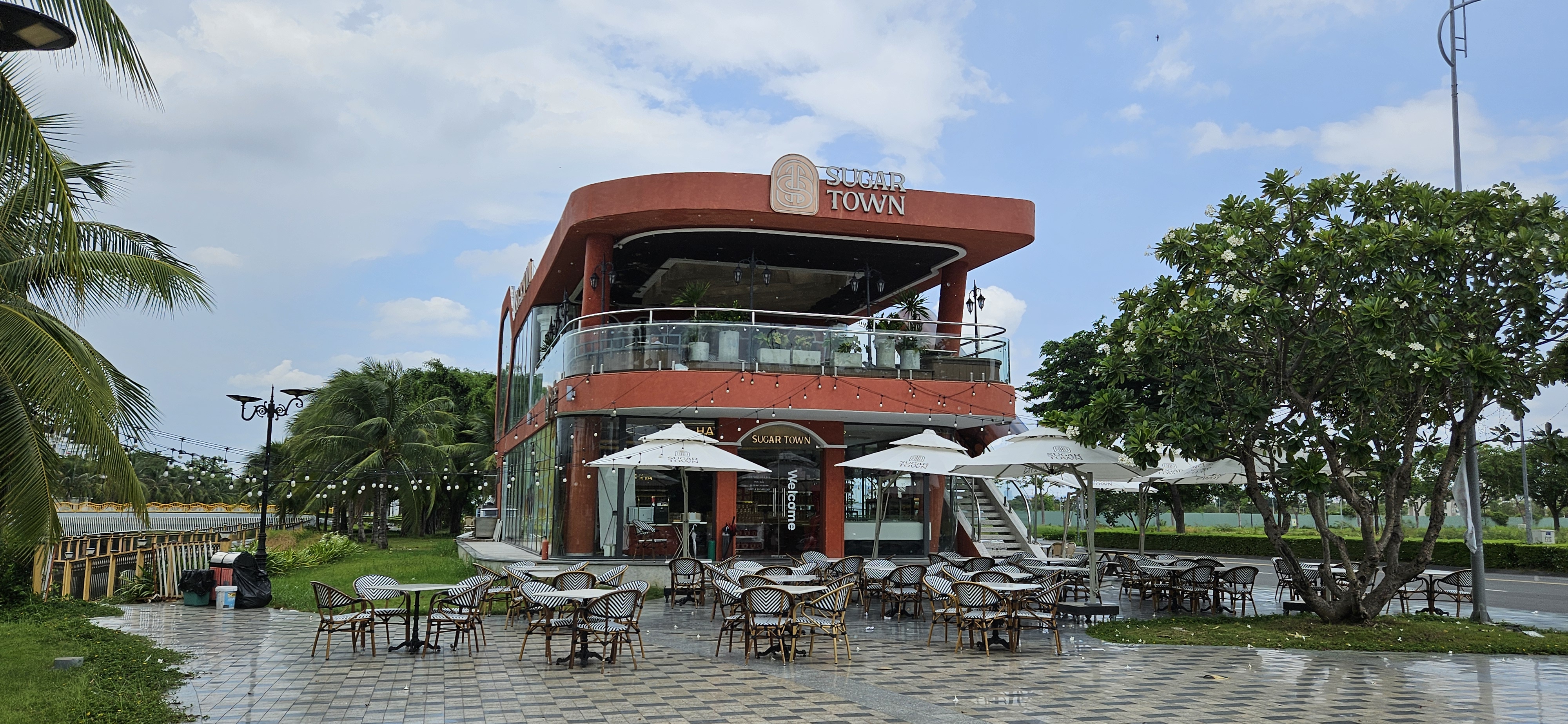 A modern restaurant named 'Sugar Town' featuring a two-story design with large glass windows, outdoor seating, and umbrellas. The surrounding area includes palm trees and a walkway lined with decorative lights.