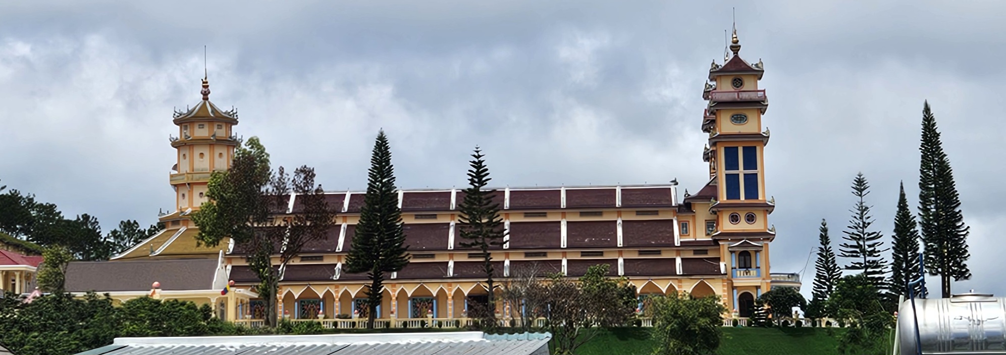 A large, ornate building with yellow and brown architecture, featuring multiple towers and surrounded by tall evergreen trees under a cloudy sky.