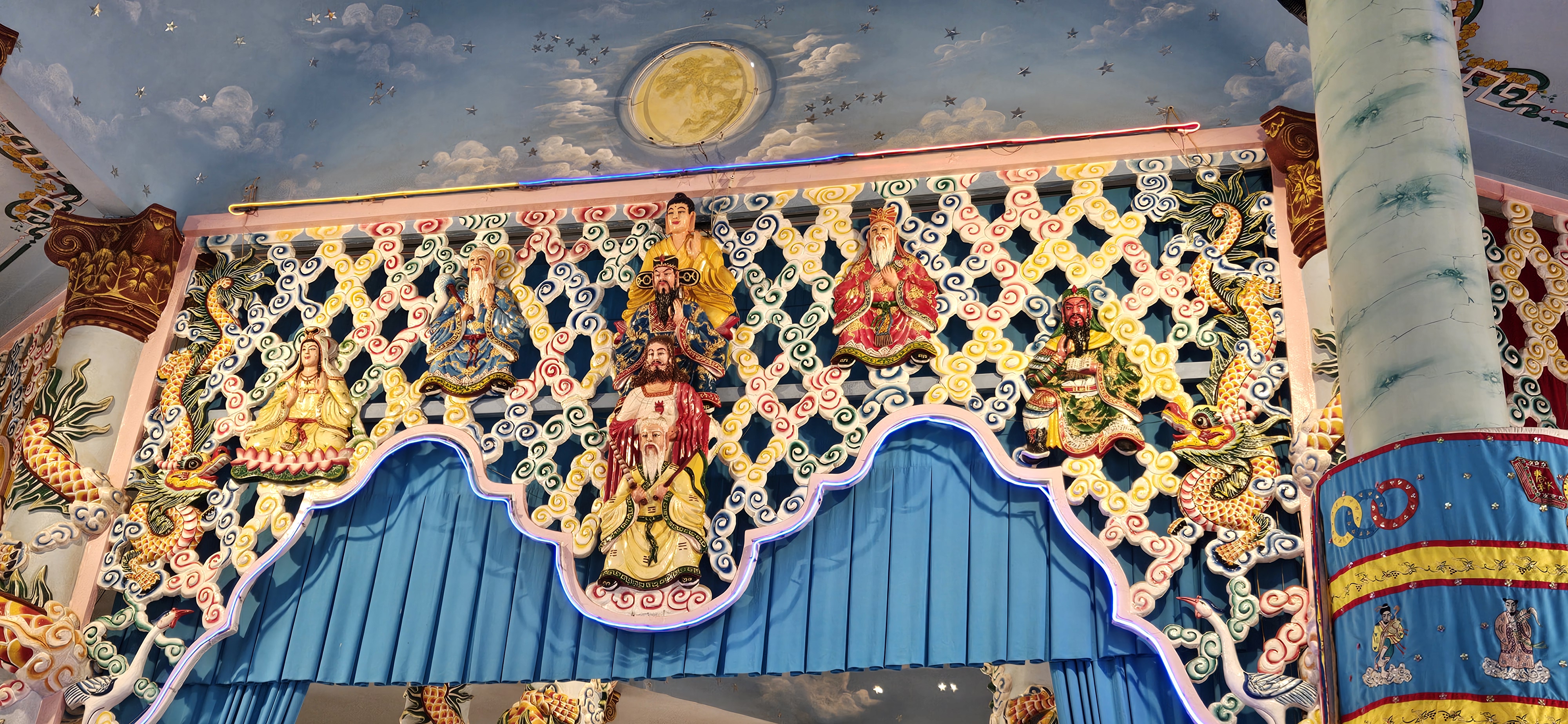 A decorative temple wall featuring colorful carvings of deities, dragons, and clouds, with intricate designs and a blue curtain at the base.