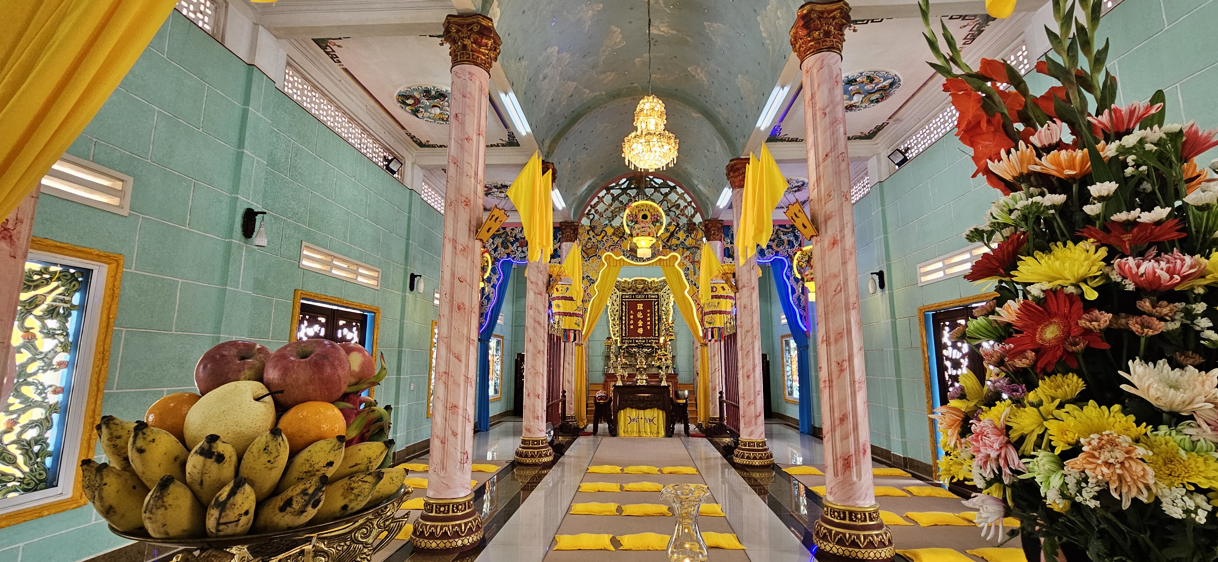 Interior of a temple with colorful decorations, marble columns, yellow drapes, and a fruit offering displayed in the foreground.