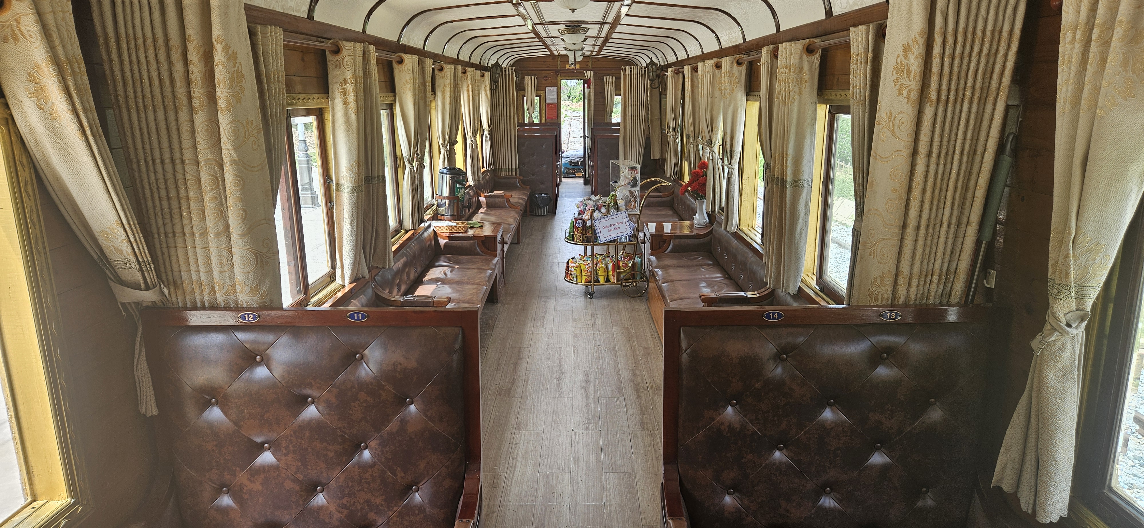 Interior of a vintage train carriage featuring elegant leather seating, large windows with curtains, and a snack cart in the aisle.