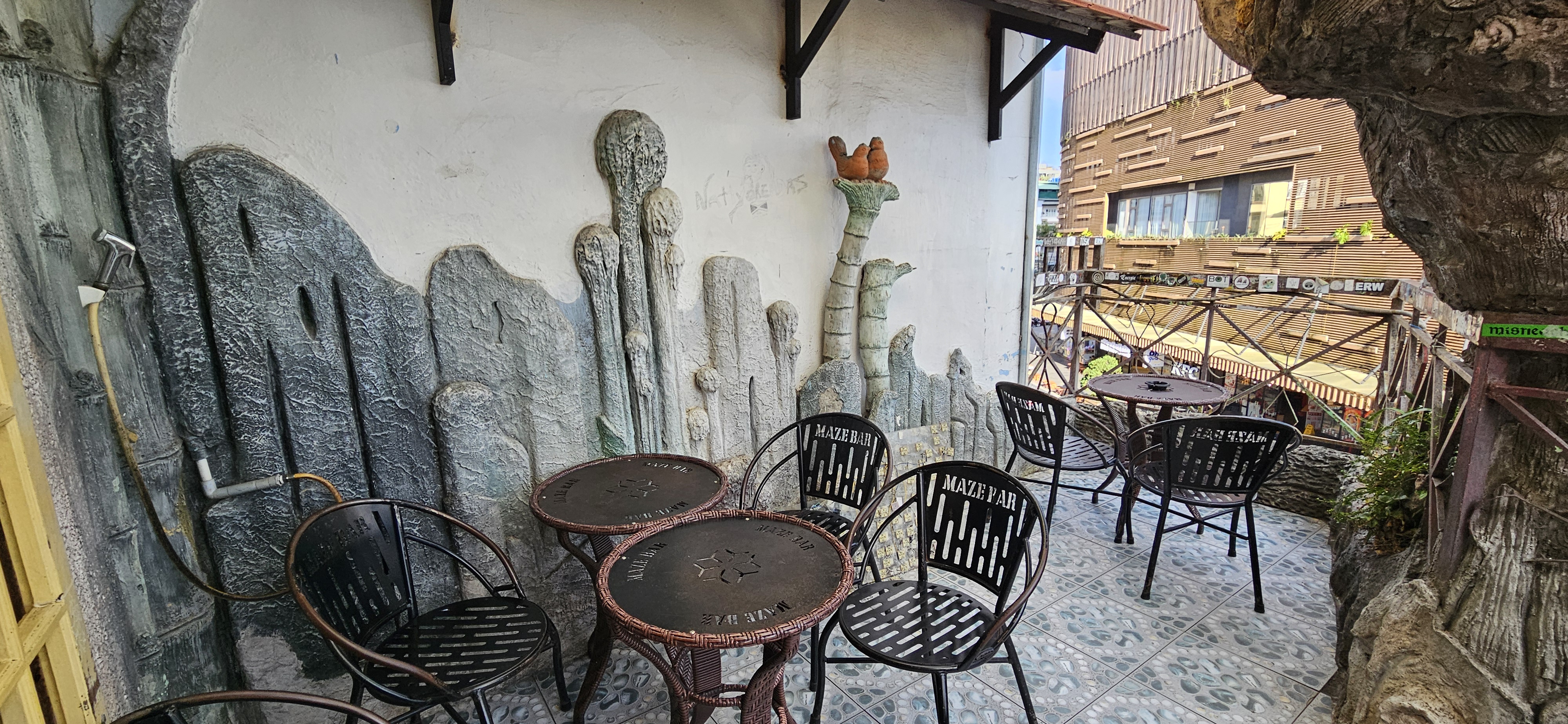 An outdoor seating area featuring black metal chairs and tables, surrounded by creatively textured walls resembling rock formations. A glimpse of the city can be seen in the background.