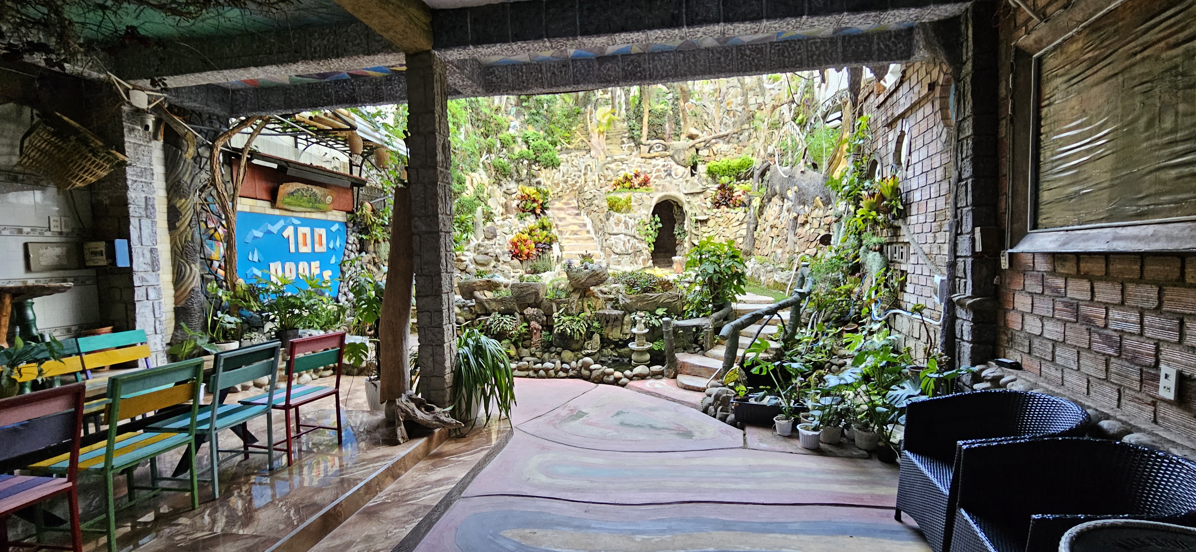Indoor garden space featuring a stone fountain, lush greenery, and colorful seating. A staircase leads to a landscaped area with plants and rock formations.