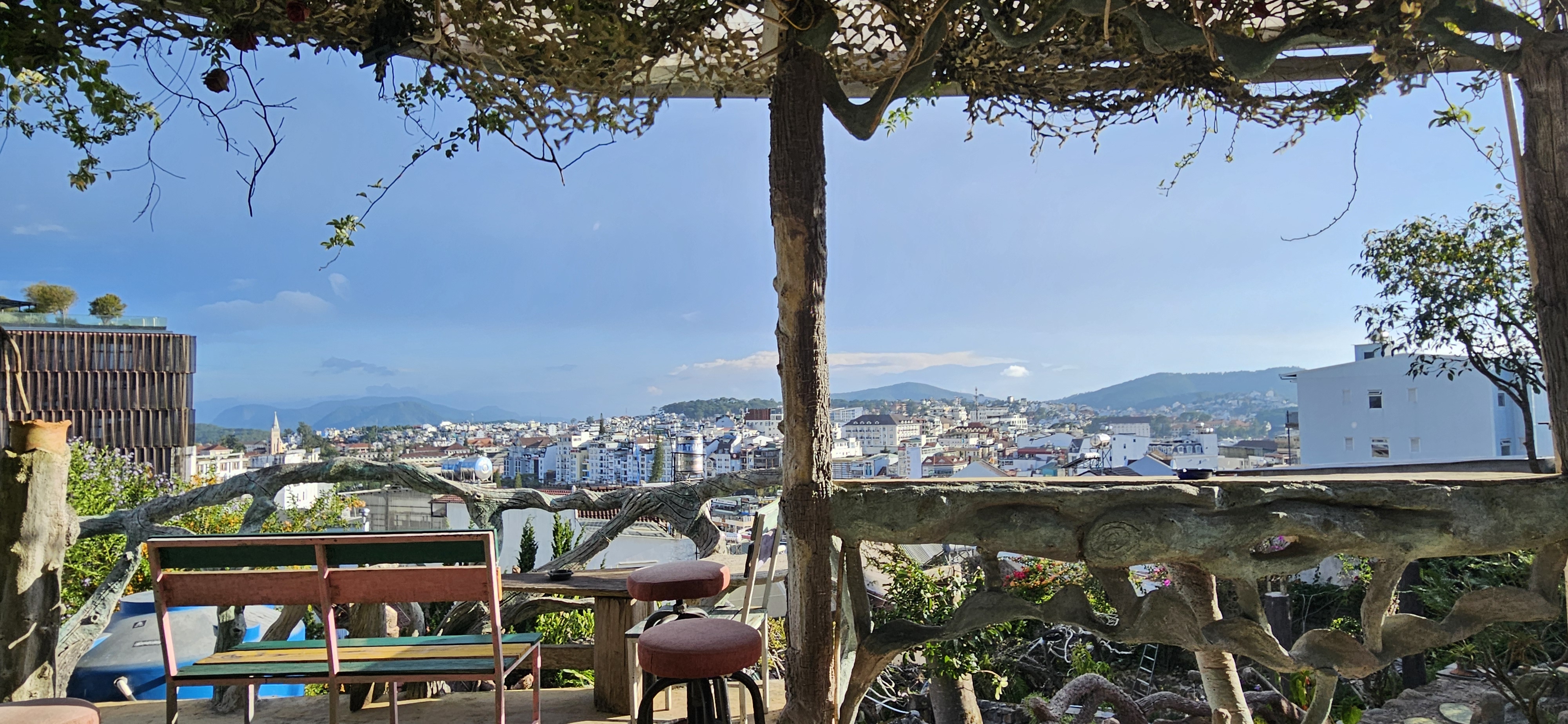 A scenic view overlooking a city from a terrace with wooden seating, framed by branches and foliage, with mountains in the background and a clear blue sky.