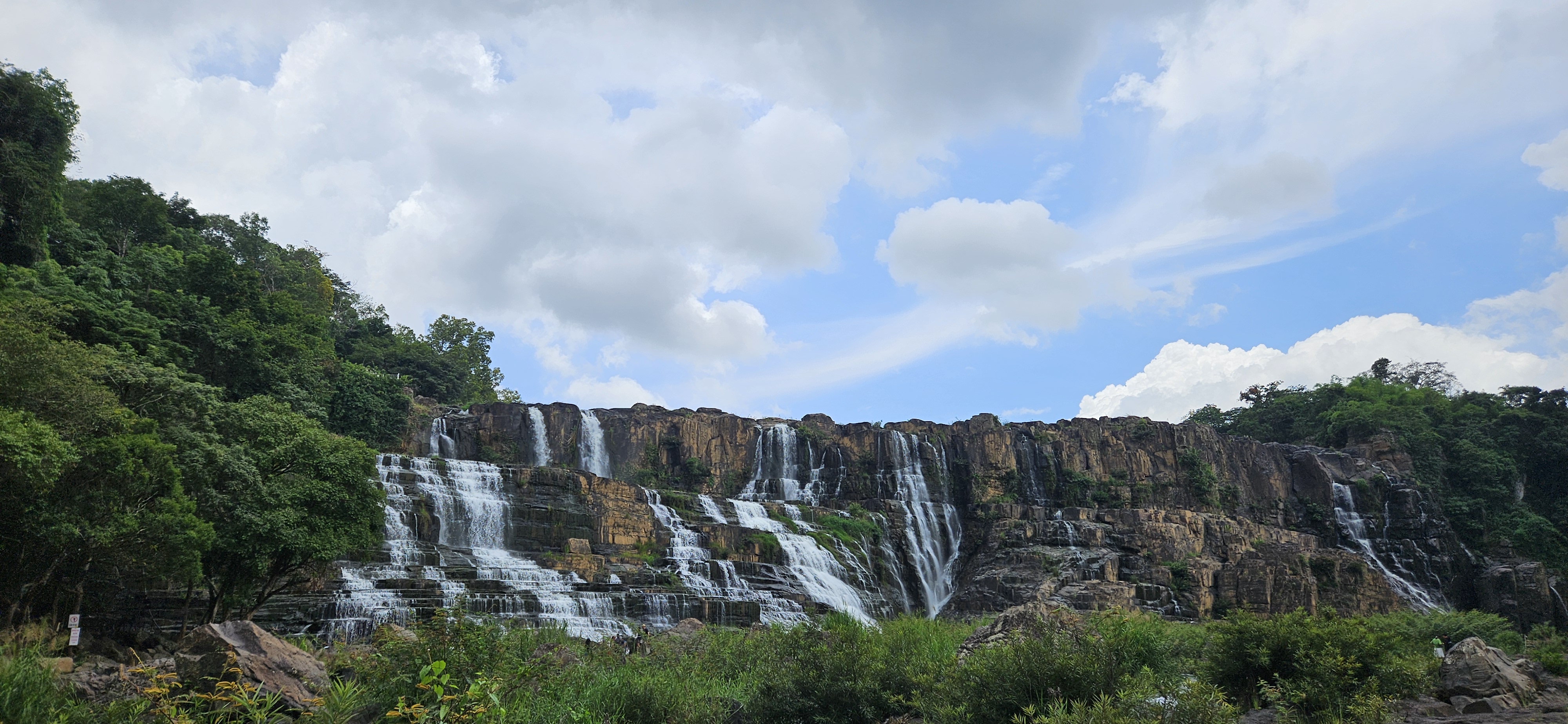 A scenic view of a waterfall cascading over rocky cliffs, surrounded by lush greenery and a partly cloudy sky.