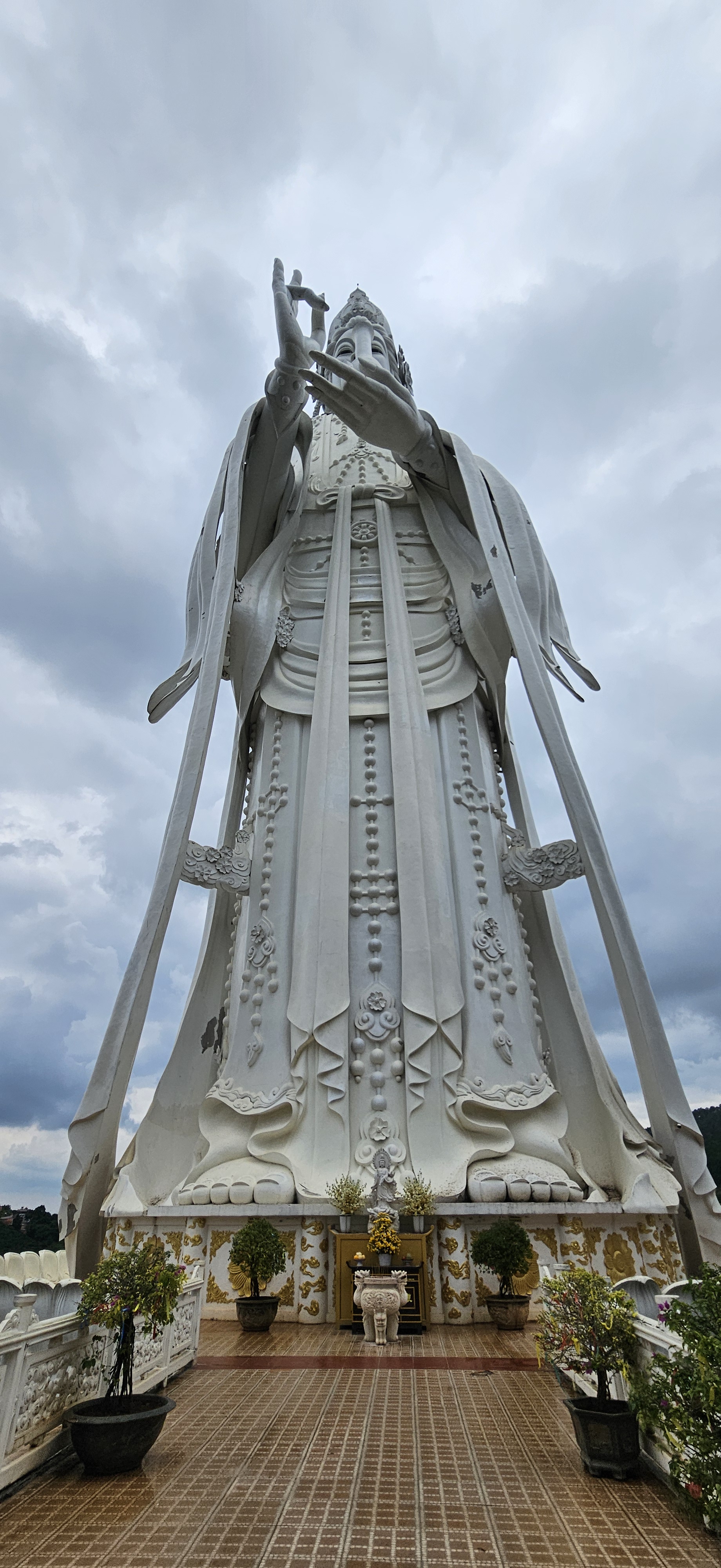 A towering white statue of a deity, viewed from below, with outstretched hands. The sky is overcast, and there are decorative plants and offerings at the base of the statue.