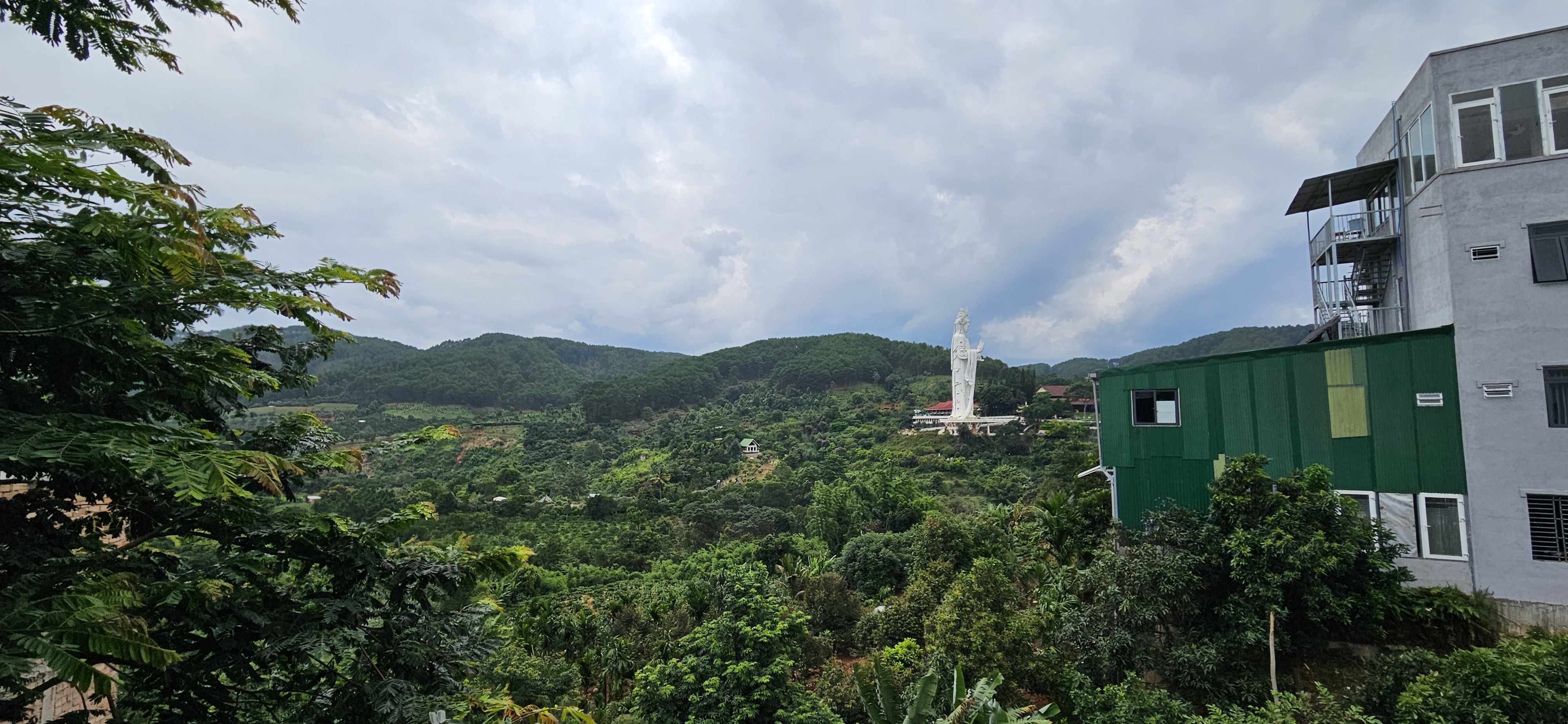 Panoramic view of lush green hills with a large white statue on the right, partially obscured by trees and residential buildings.