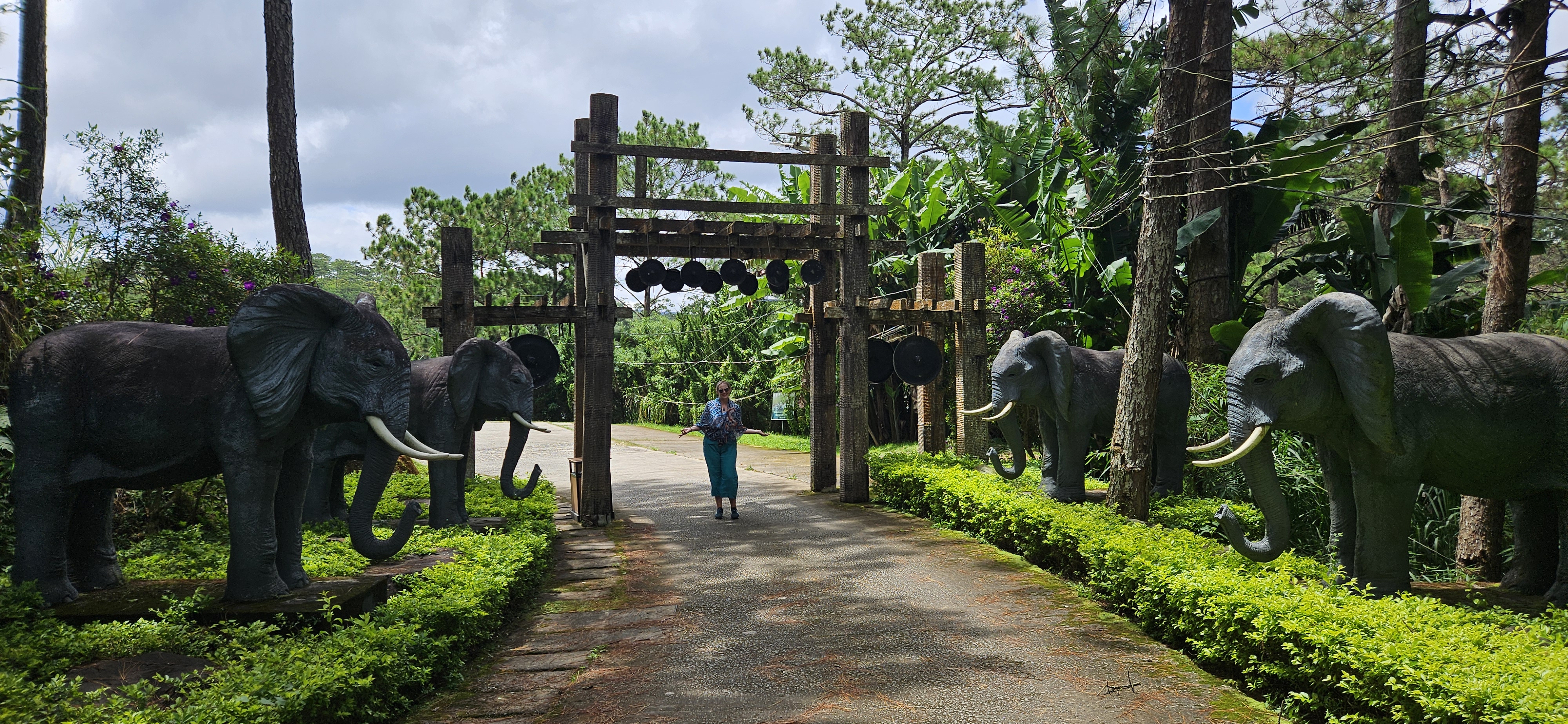 A person standing in front of a wooden archway with two elephant statues on either side, surrounded by lush greenery and trees.