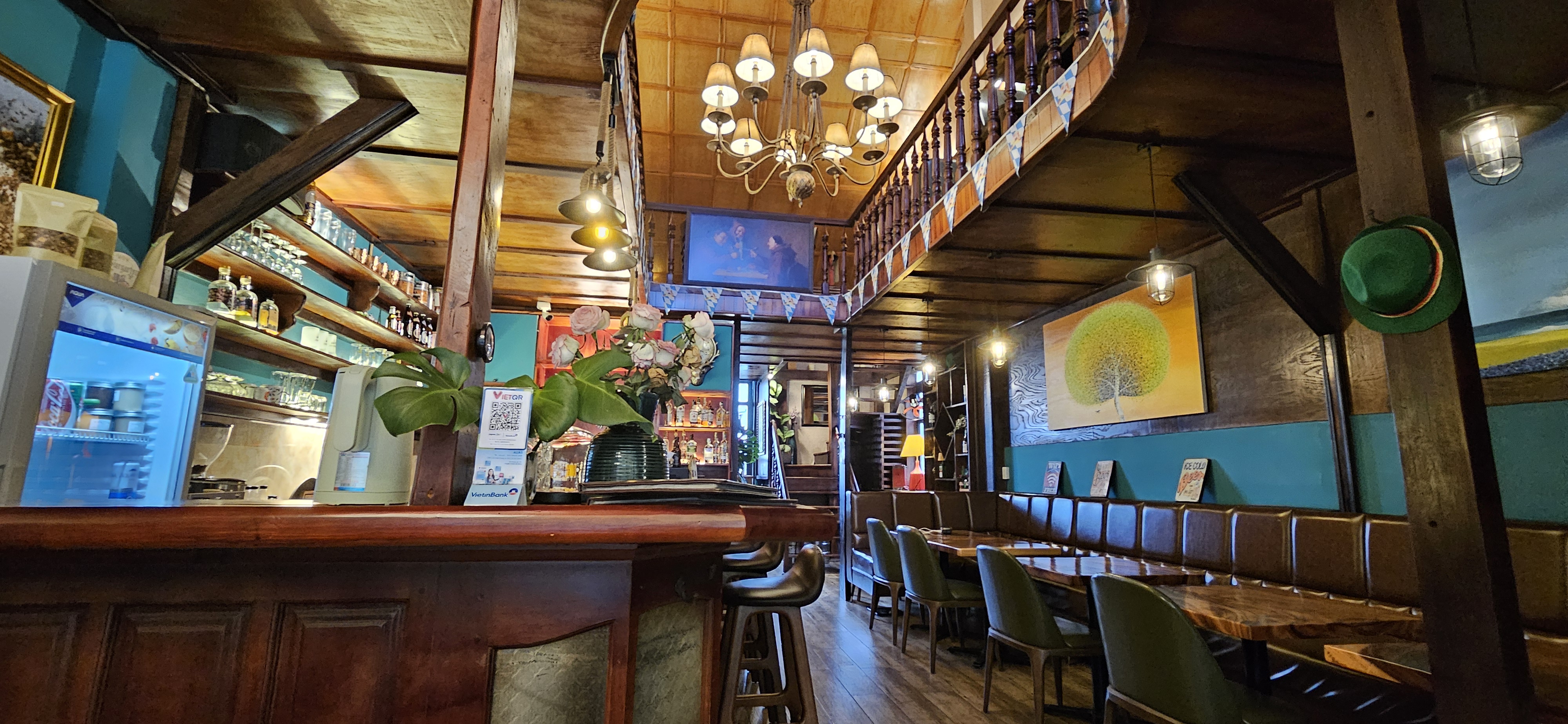 Interior view of a cozy cafe featuring wooden decor, a large chandelier, and a well-stocked bar. There are tables and chairs arranged on the floor, with a staircase leading to an upper level. Various artworks adorn the walls, and potted plants add a touch of greenery.