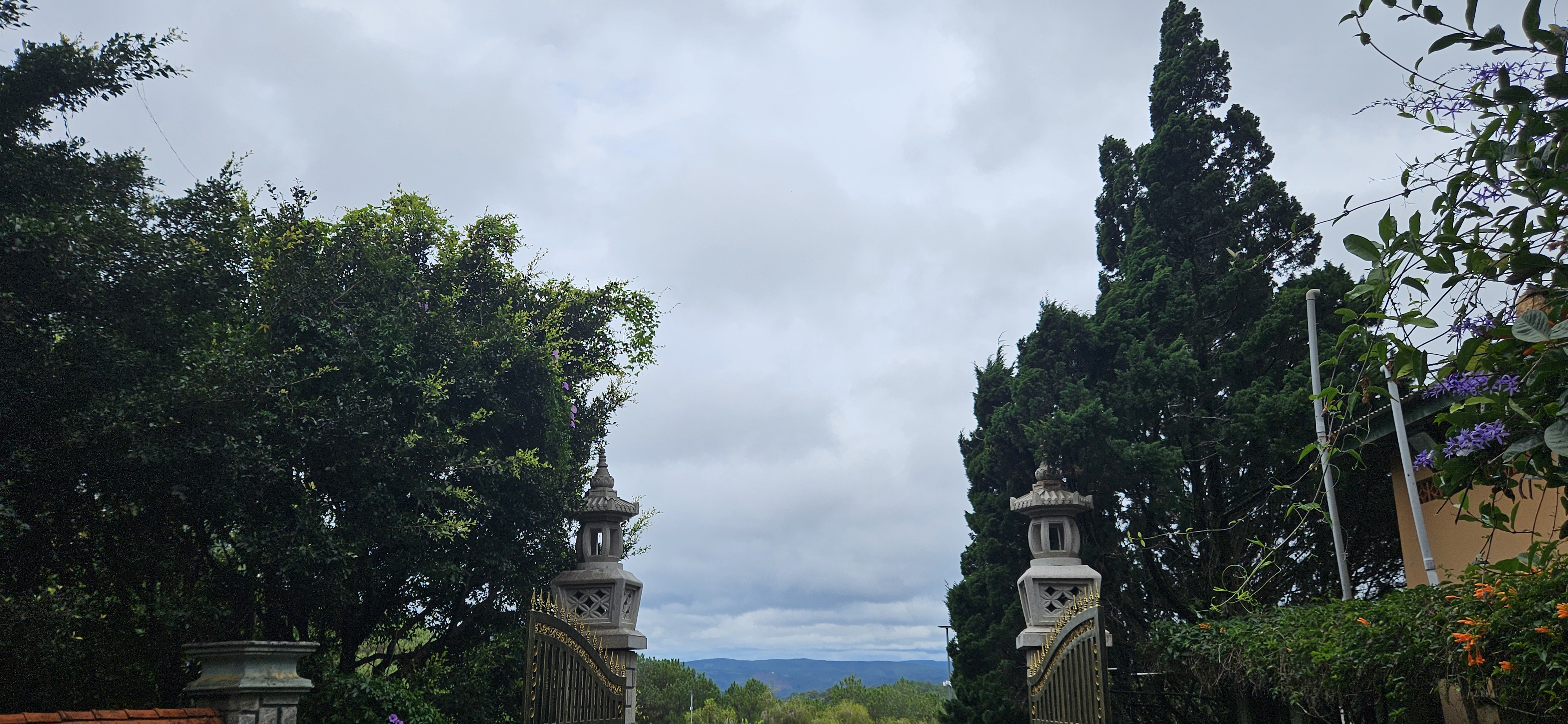 A scenic view through an ornate stone gate framed by lush green trees, with a cloudy sky and distant mountains in the background.
