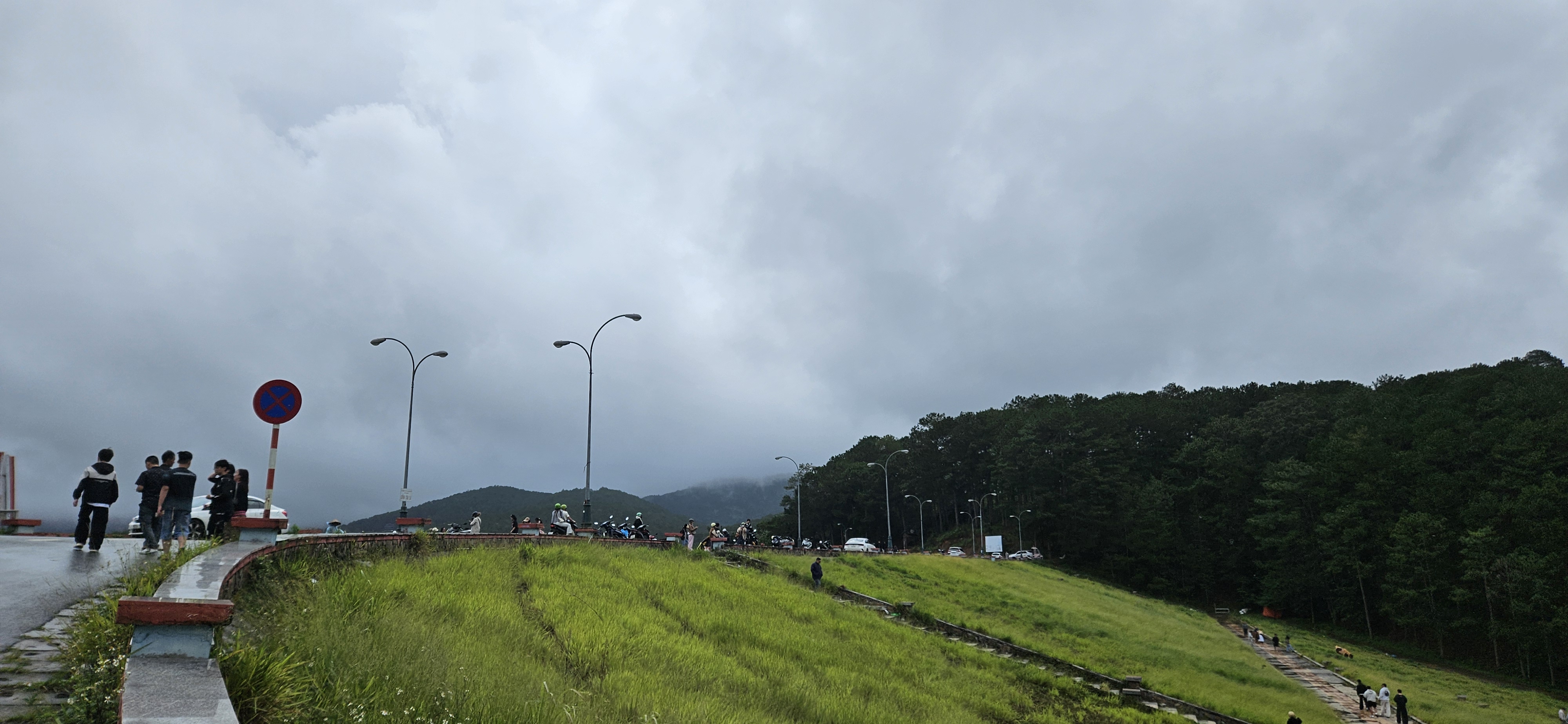 A group of people standing near a curved road with green grass and trees on the right, under a cloudy sky.