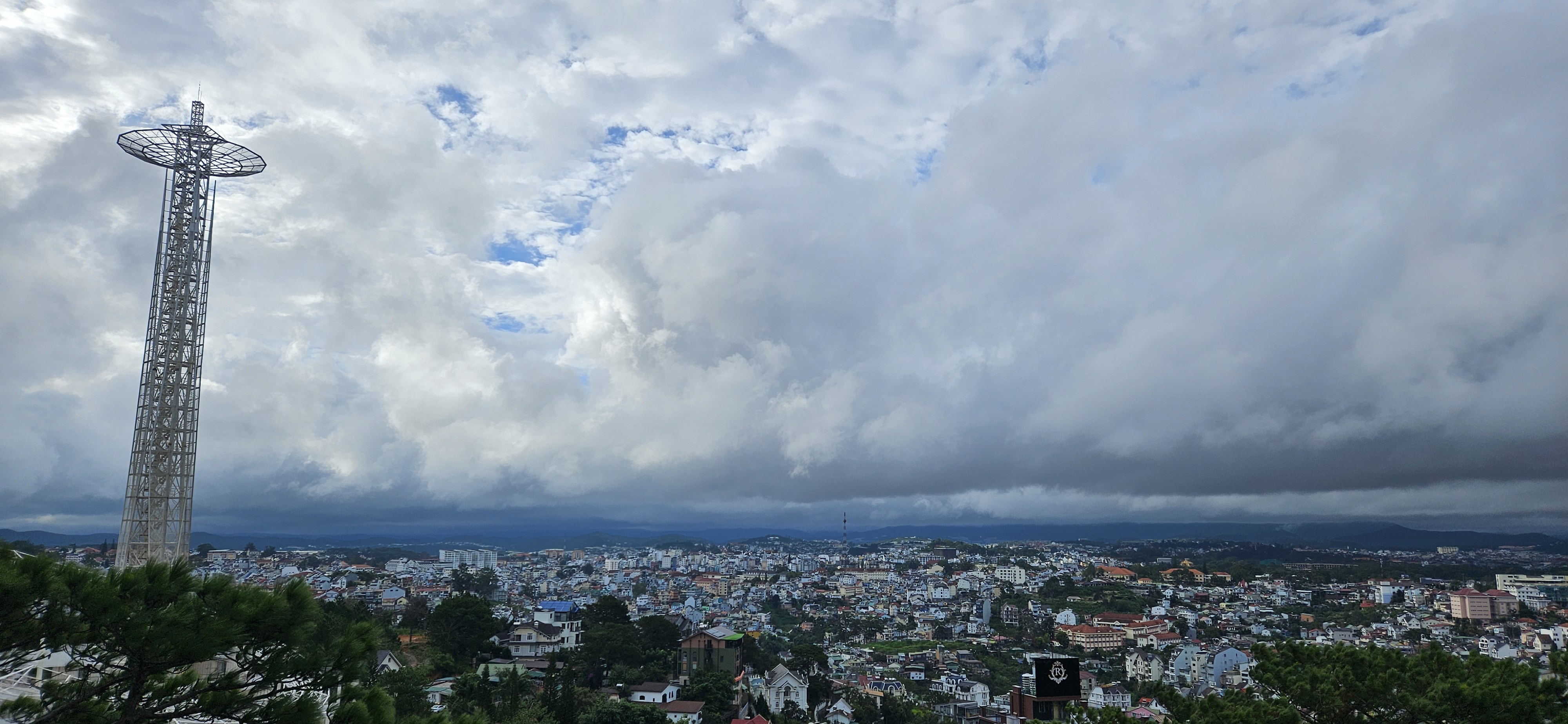 Panoramic view of a cityscape under cloudy skies, featuring a tall communication tower and a mix of residential buildings surrounded by green trees.