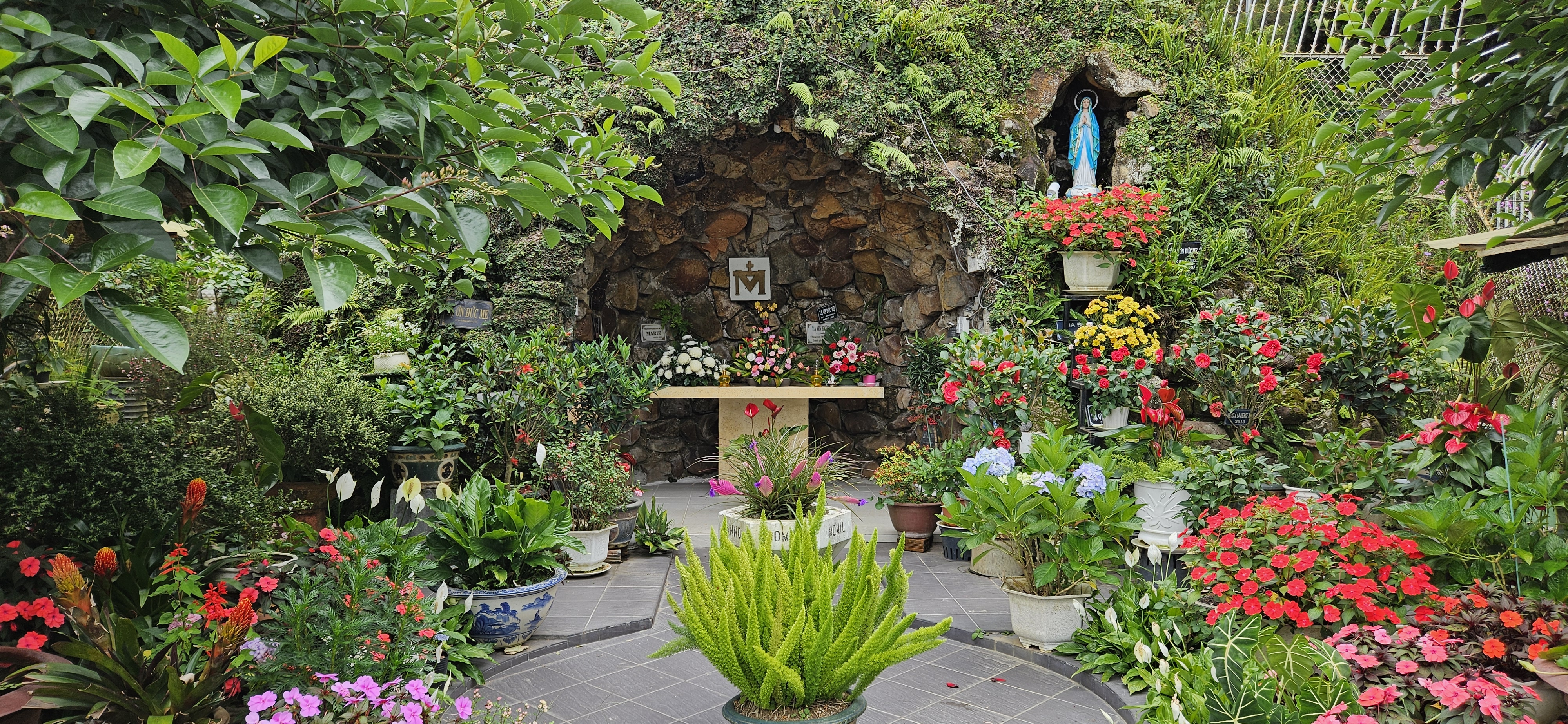 A serene garden with vibrant flowers and lush greenery surrounding a stone altar featuring a religious symbol and a statue of Mary.