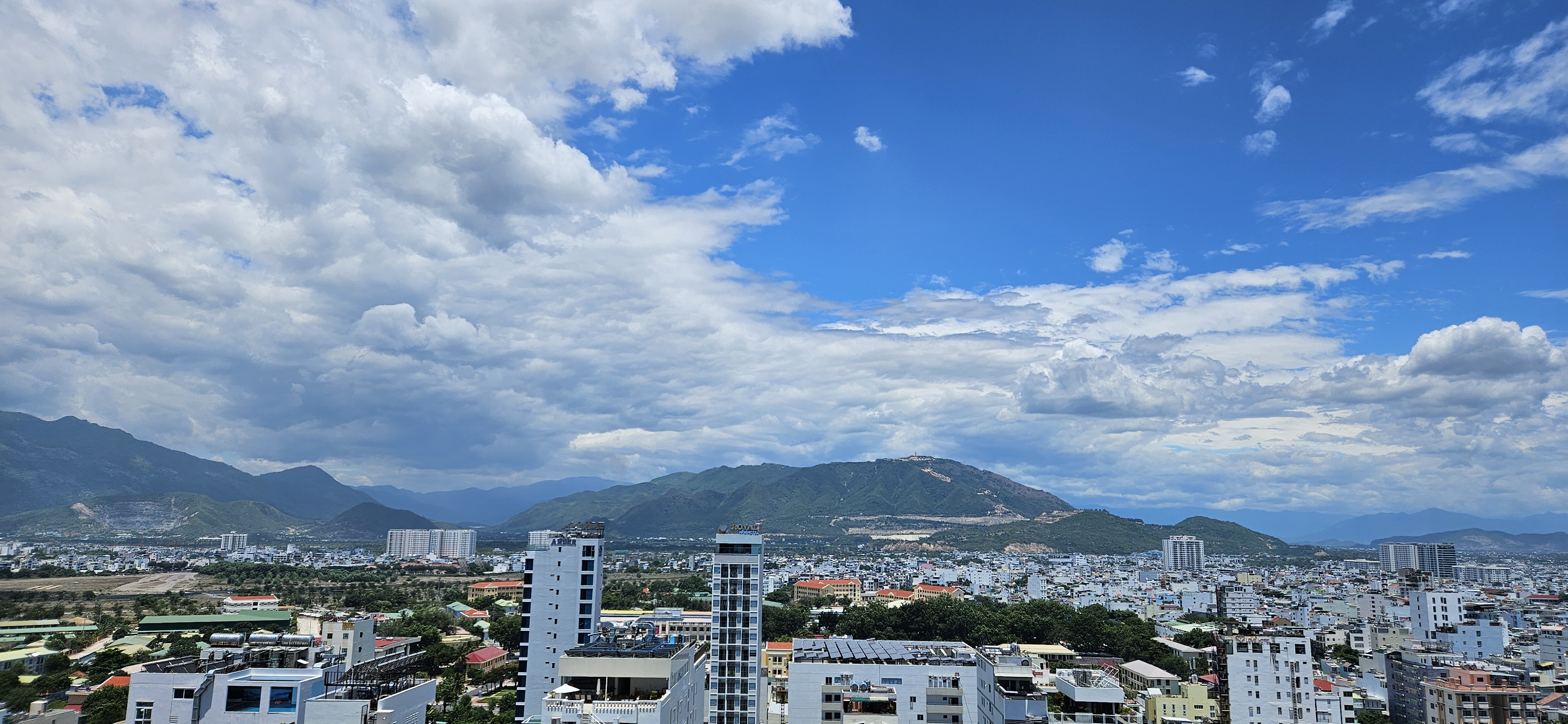 Panoramic view of a cityscape featuring buildings and mountains under a partly cloudy sky.
