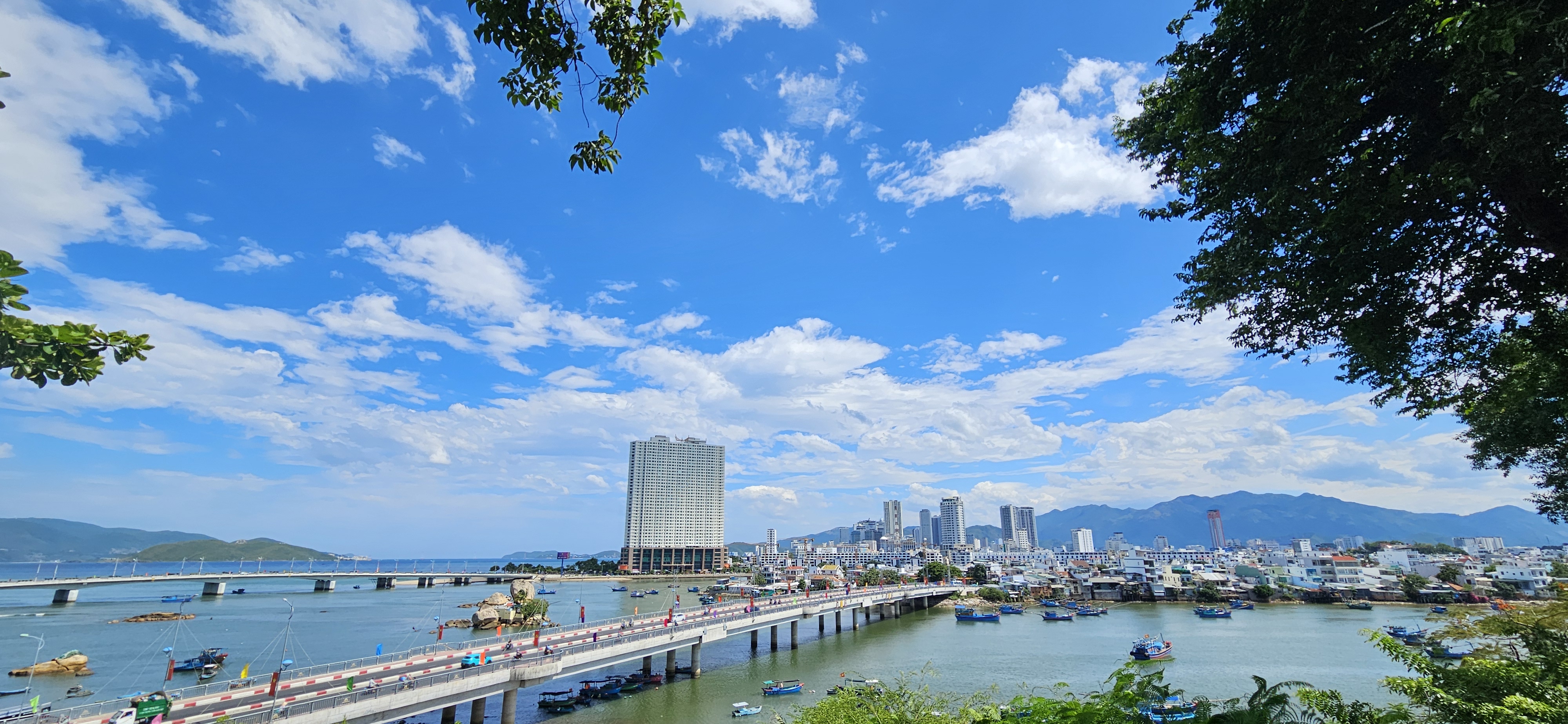 Scenic view of a cityscape by the water, featuring a bridge, a mix of modern buildings, and boats on the river under a blue sky with clouds.