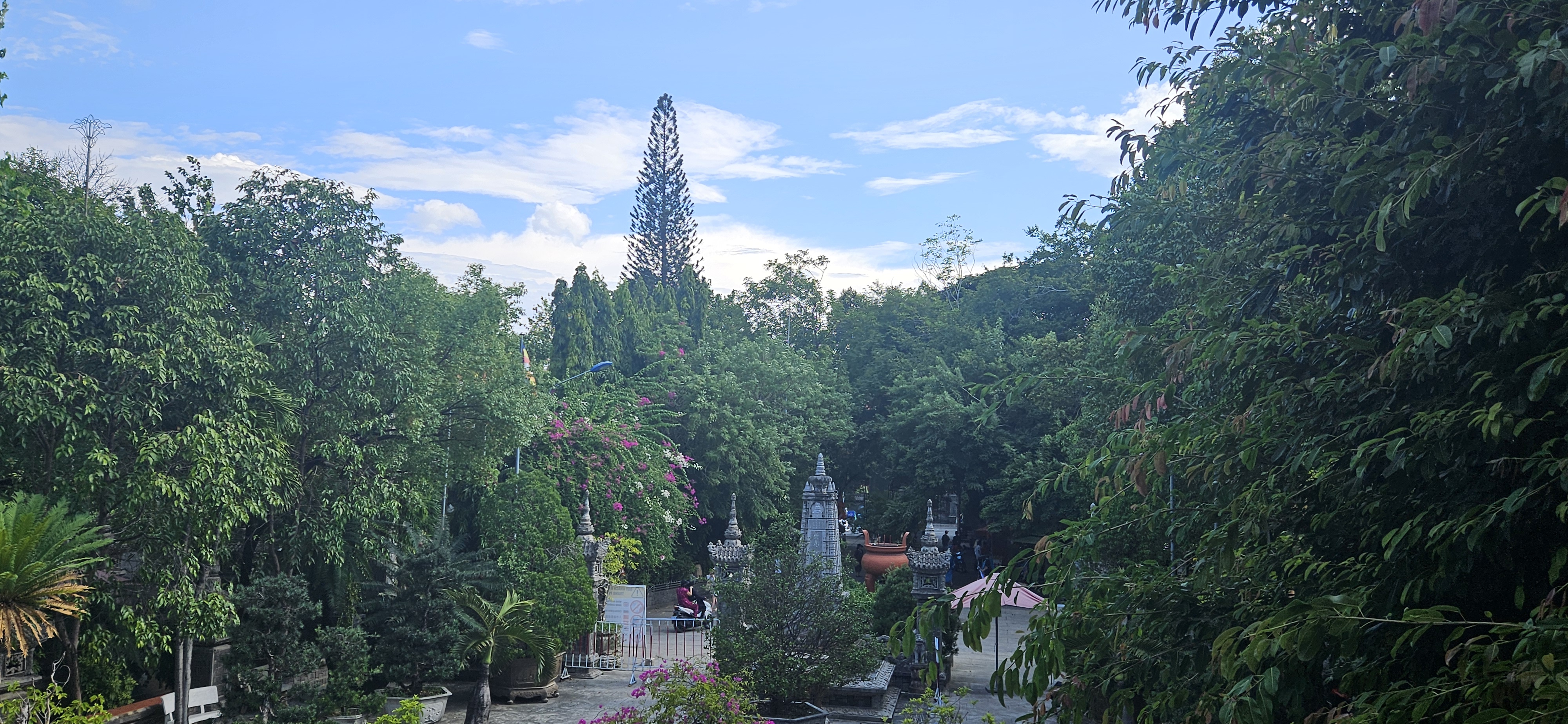 A serene garden landscape with lush green trees, colorful flowers, and a clear blue sky, featuring decorative stone structures and a glimpse of visitors enjoying the peaceful setting.