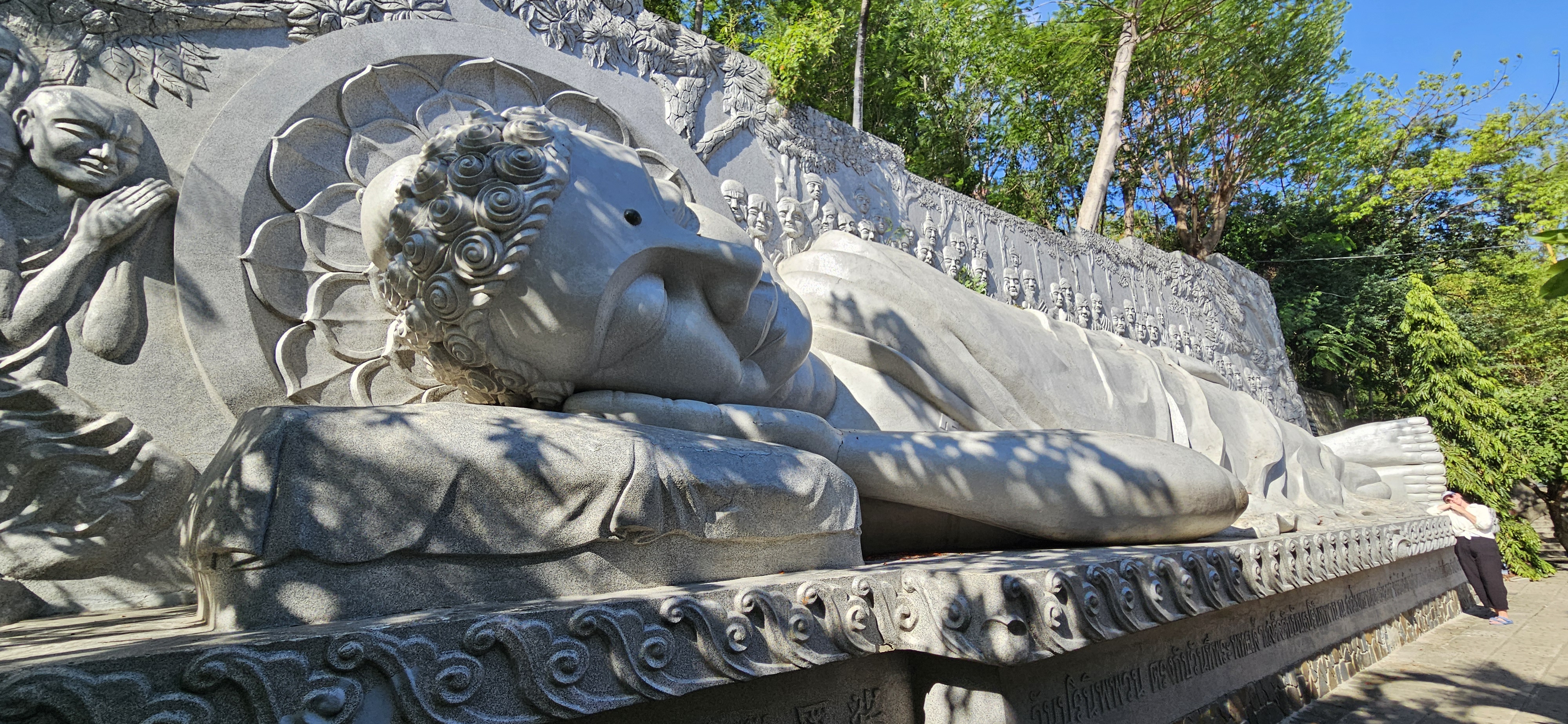 A large reclining Buddha statue made of stone, surrounded by detailed carvings and lush greenery.