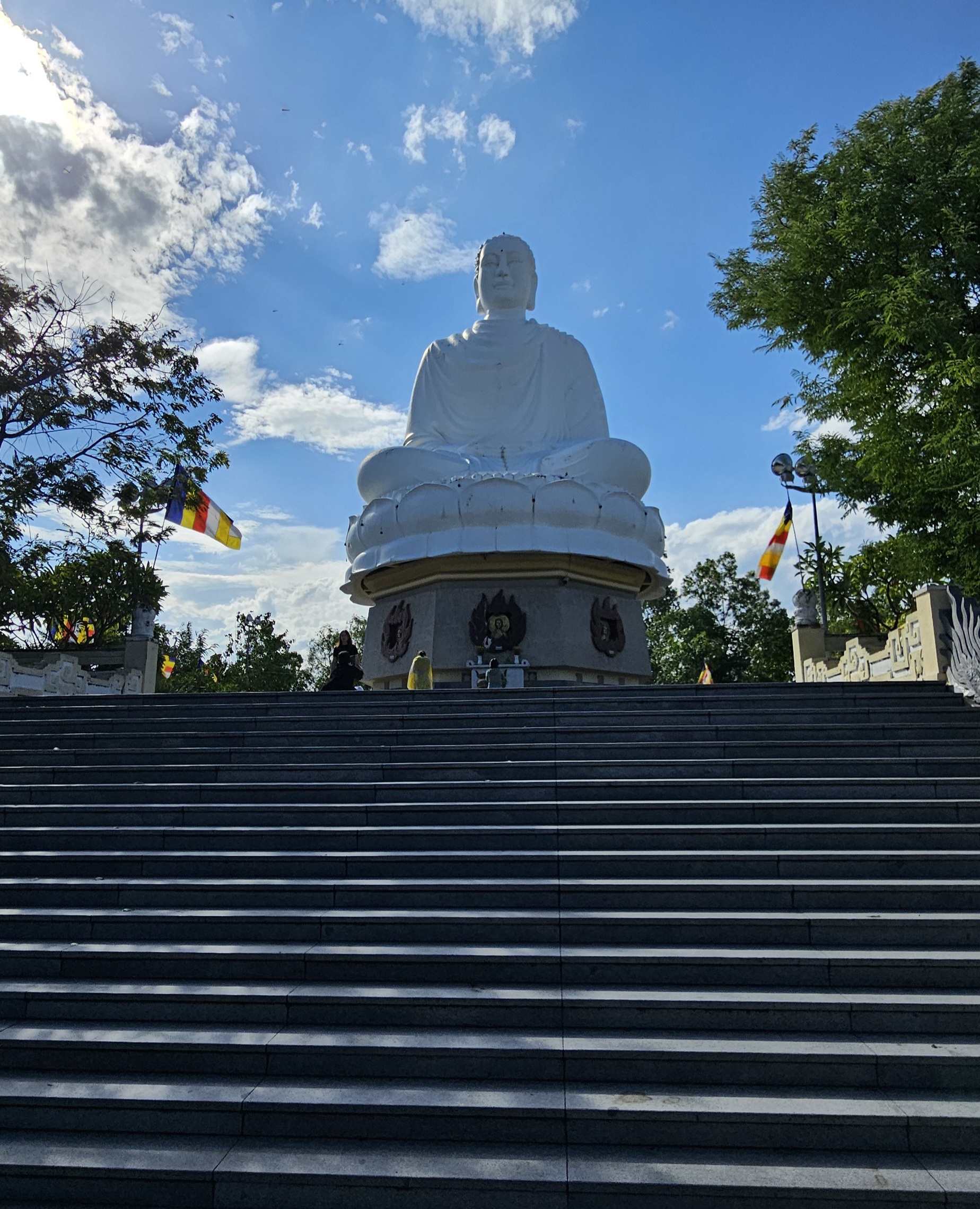 A large white statue of Buddha seated on a lotus pedestal, surrounded by green trees and colorful flags under a blue sky with clouds.