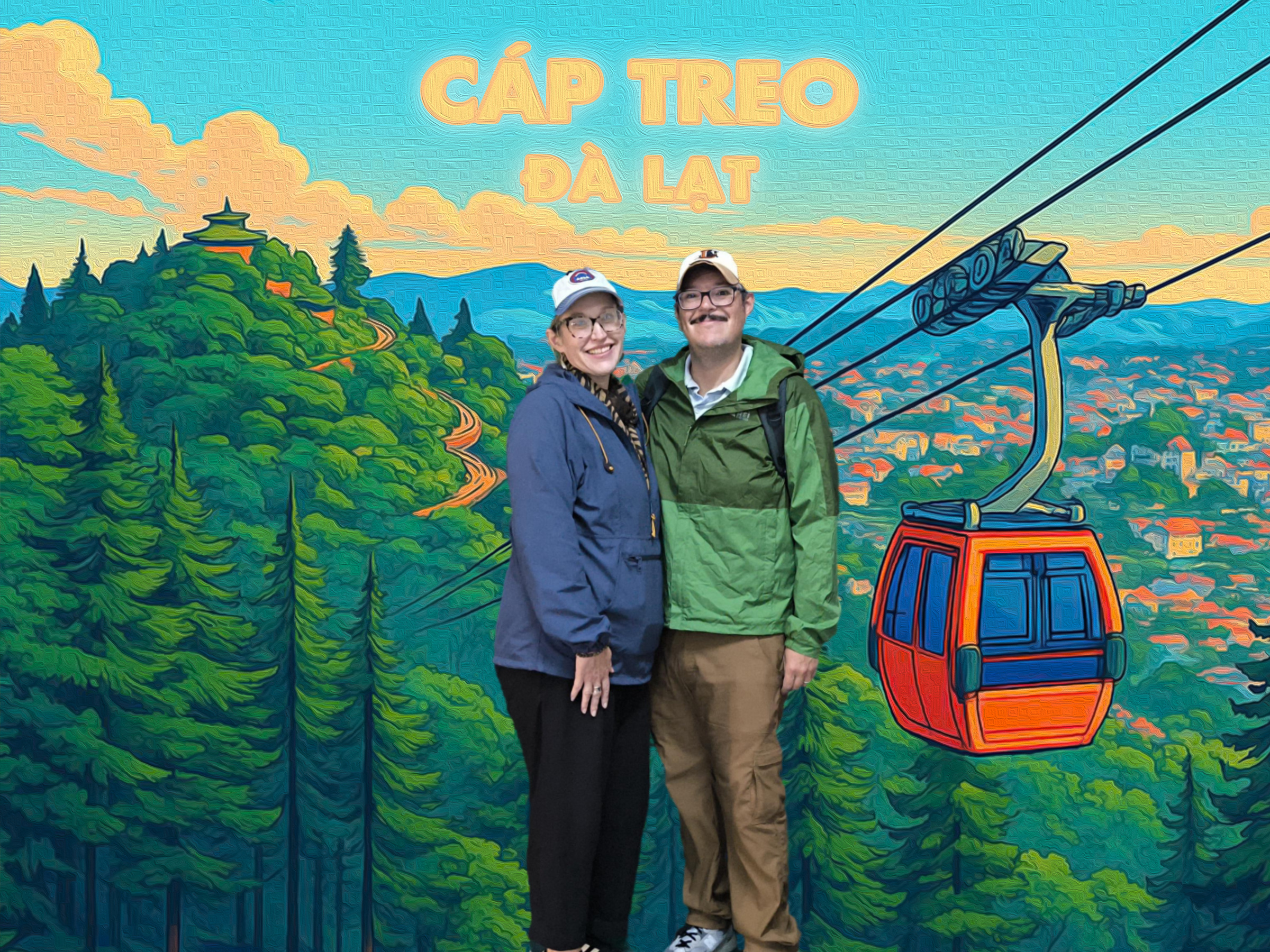 A couple poses in front of a colorful mural depicting a mountain landscape with a cable car and lush greenery, featuring the words 'Cáp Treo Đà Lạt'.