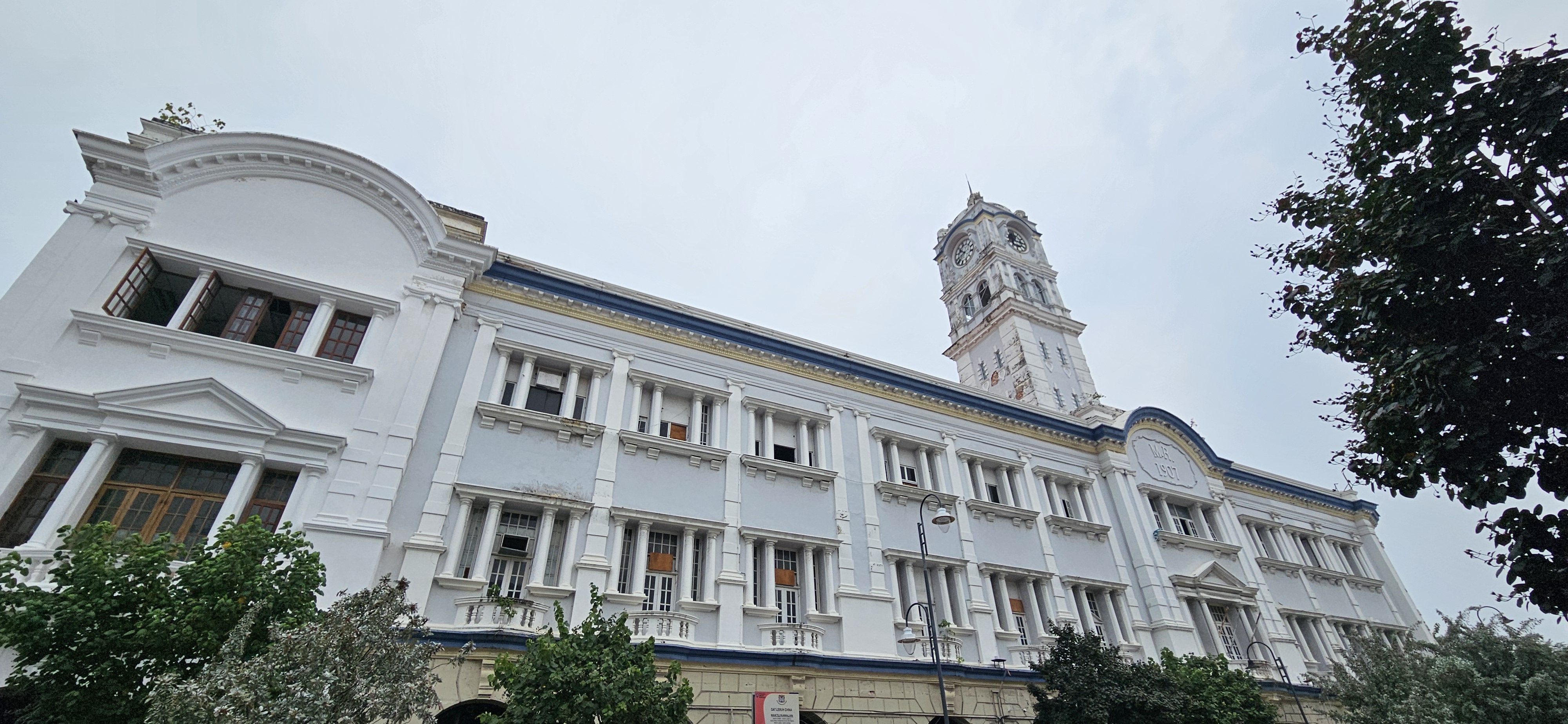 Historic white building with a clock tower, featuring classical architectural details and green foliage in the foreground.