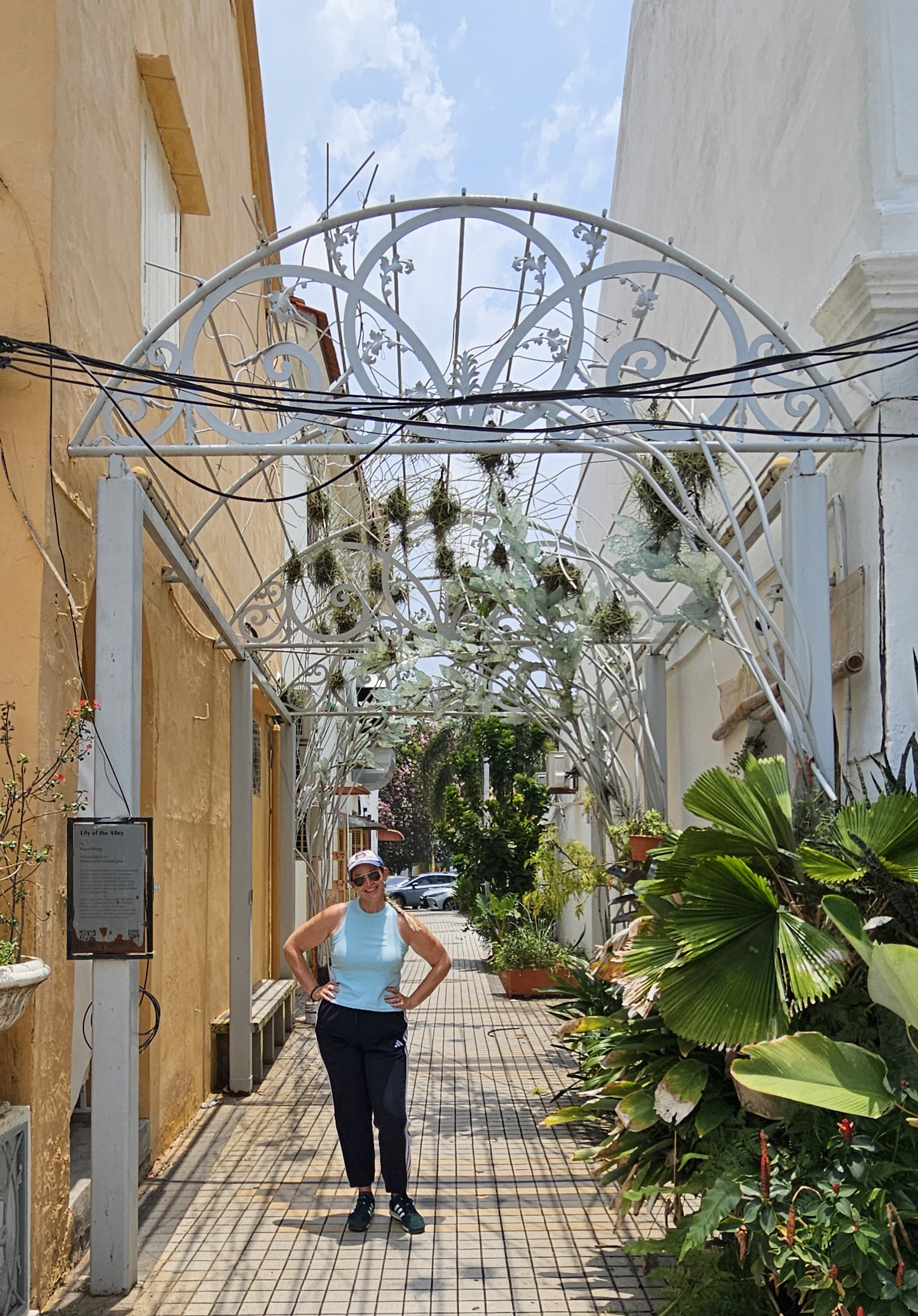 A woman standing under a decorative archway in a sunny alley, surrounded by greenery and colorful plants.