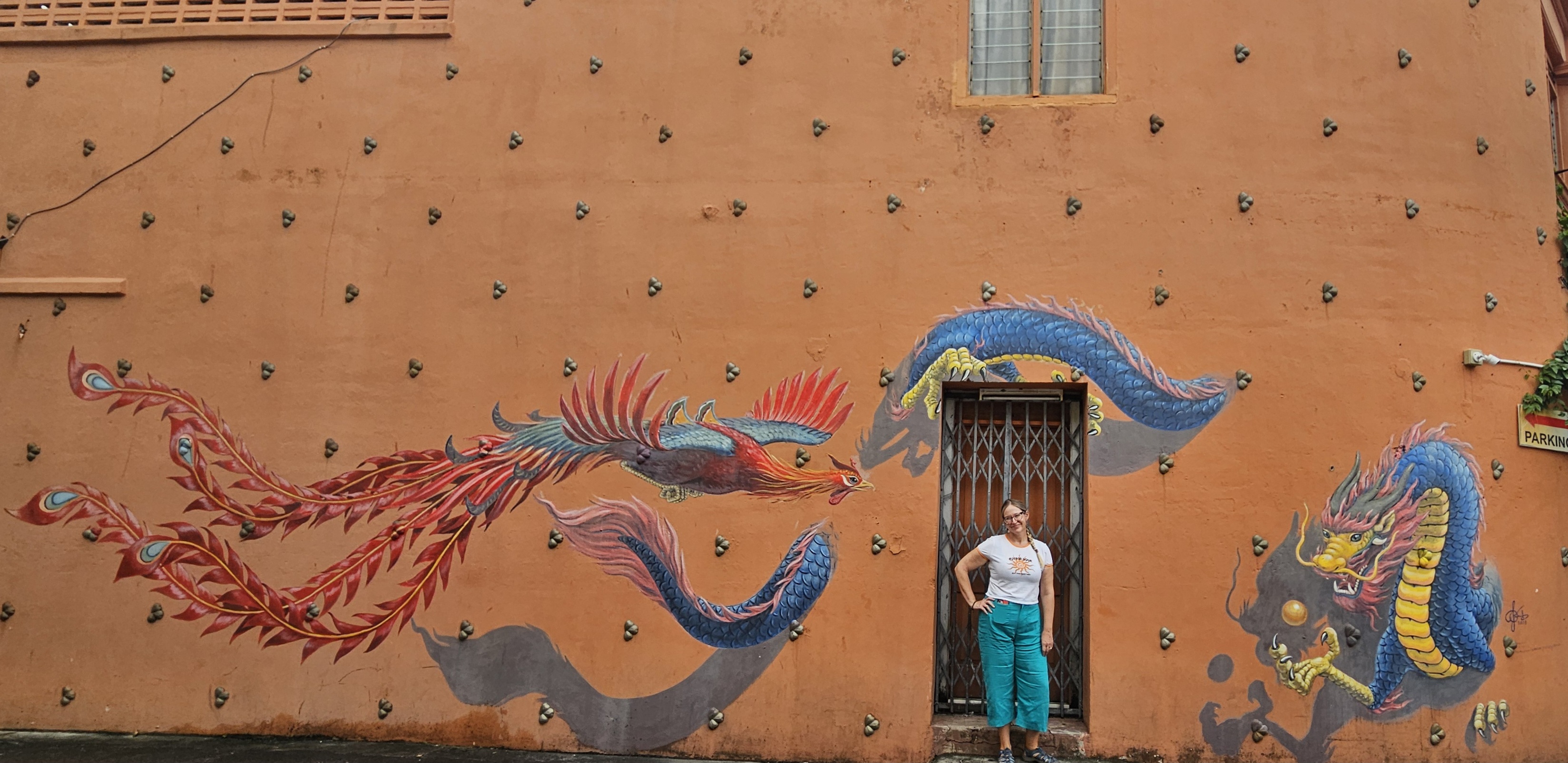 A mural on an orange wall featuring a colorful dragon and a phoenix, with a person standing in front of it.