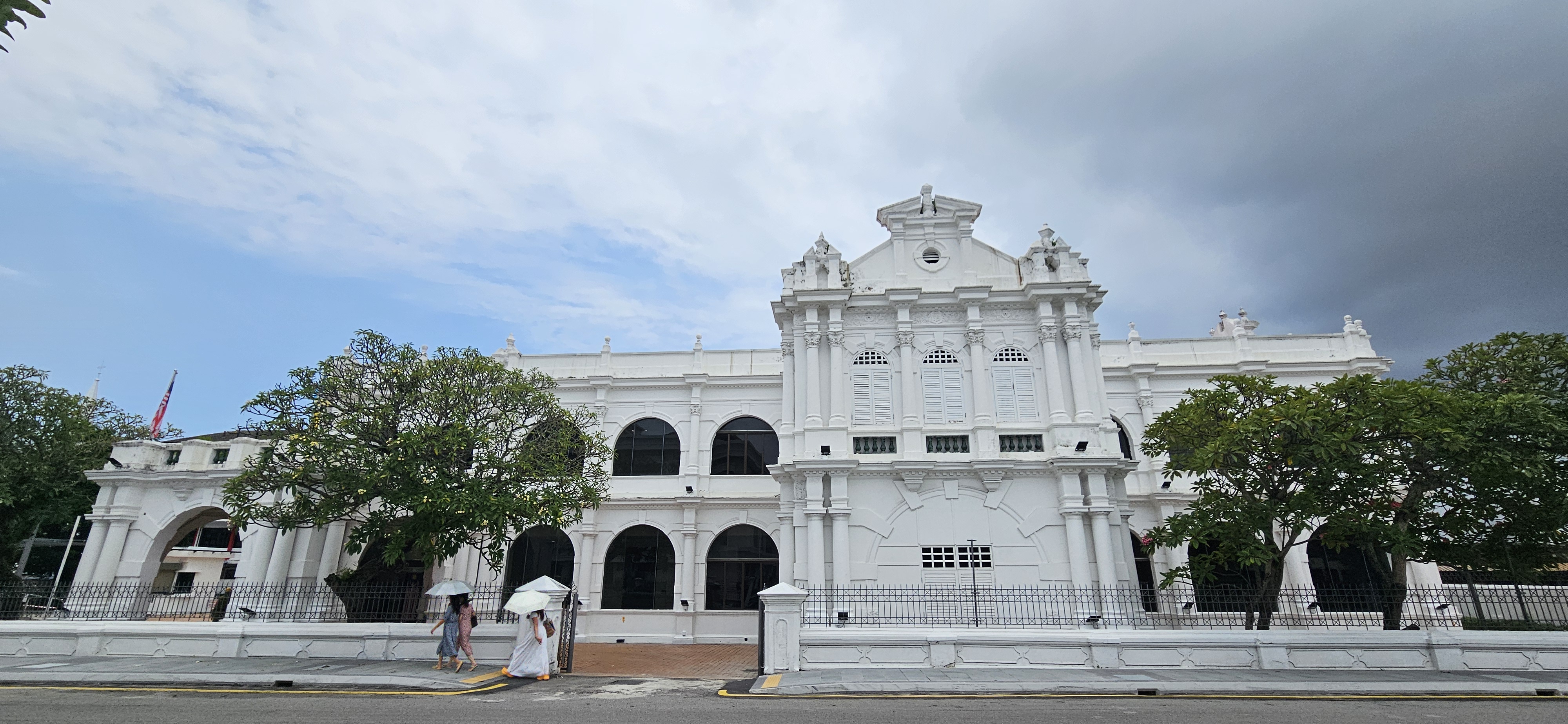 A historic white building with ornate architecture, featuring large arched windows and a decorative façade, surrounded by greenery, under a partly cloudy sky.