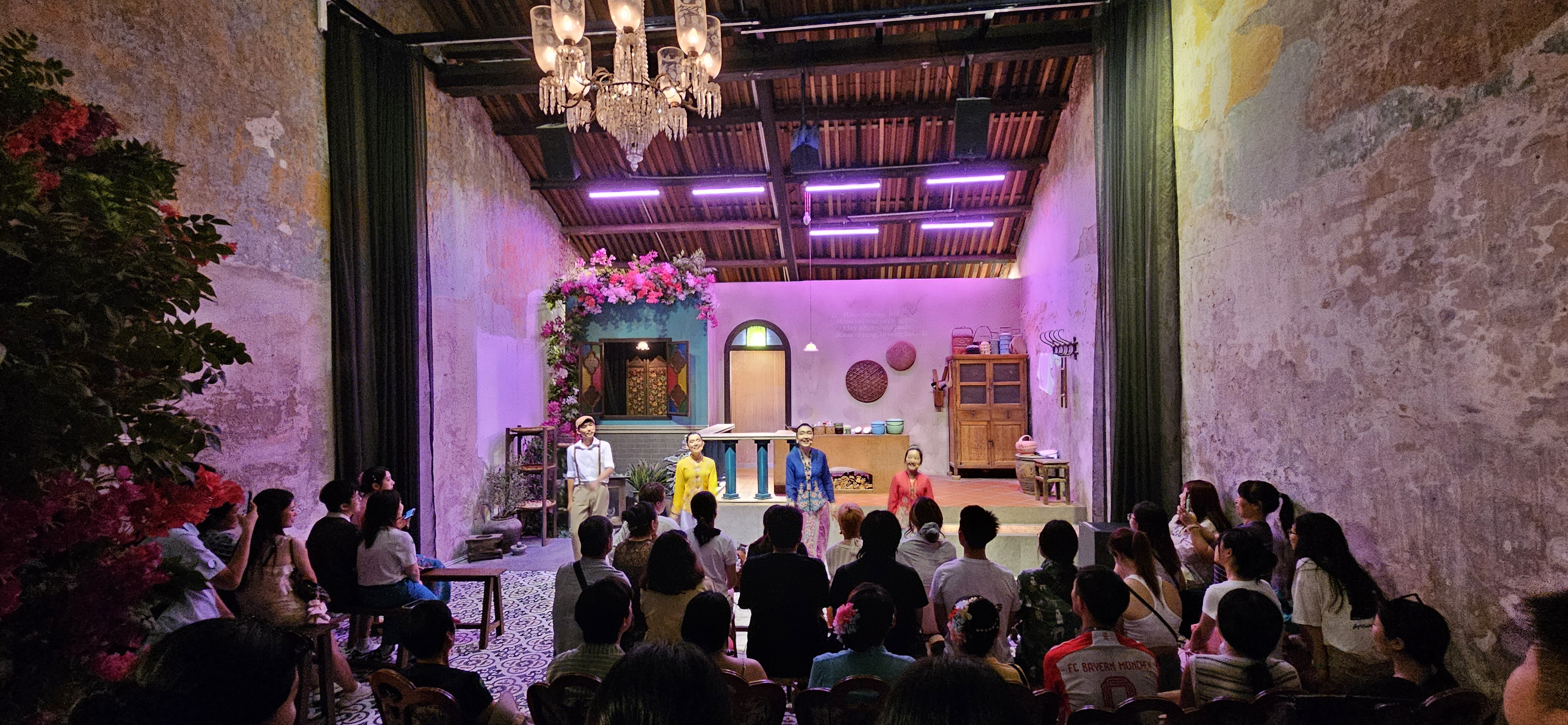 A brightly colored cultural performance taking place in a vintage-styled venue with a lively audience. Performing artists are on stage, surrounded by floral decor and purple lighting, while a diverse crowd watches attentively.